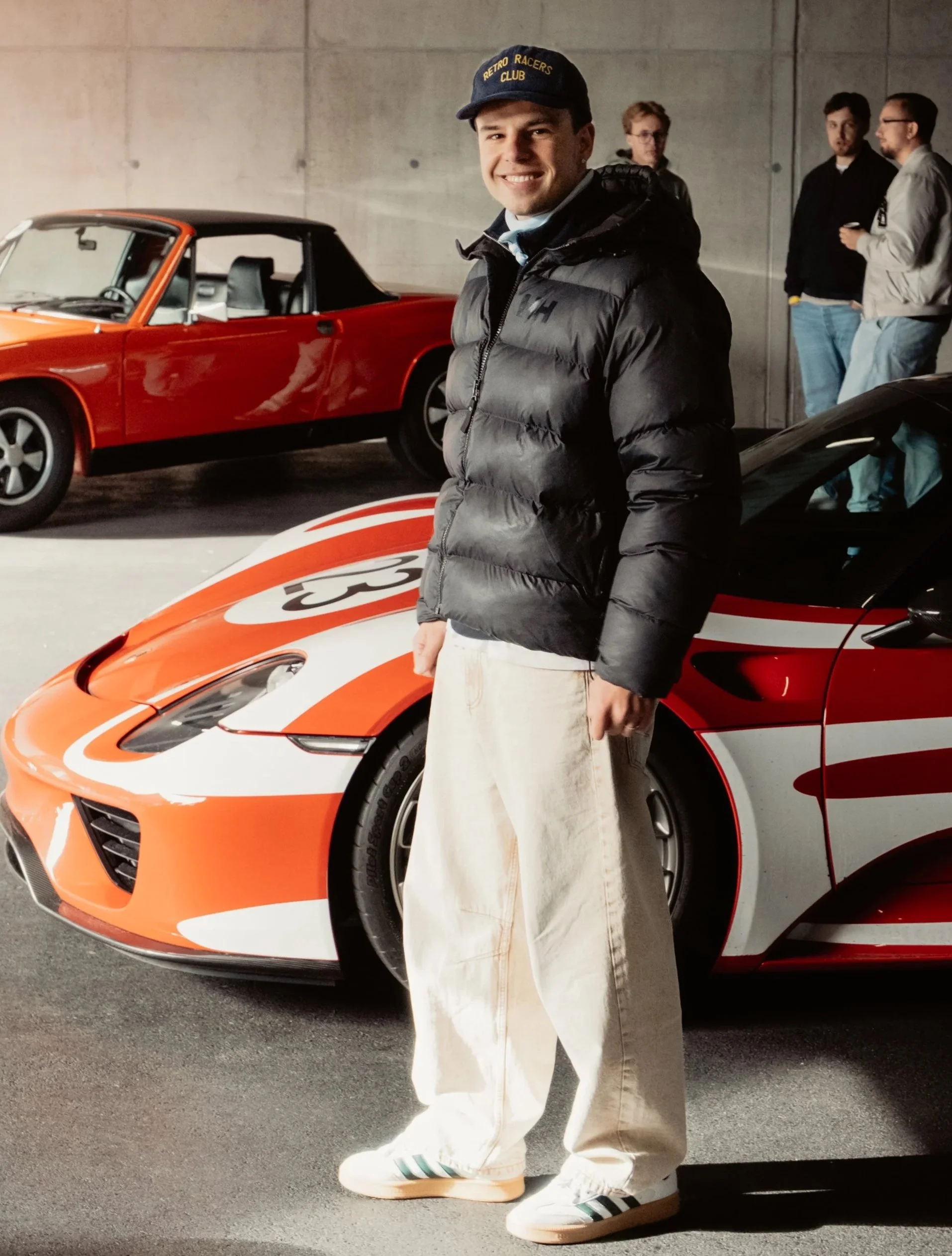 Junger Mann steht vor einem rot-weißen Sportwagen in einer Garage, trägt schwarze Jacke, helle Jeans und einen schwarzen Cap mit gelbem Text, im Hintergrund stehen drei Männer neben einem roten Oldtimer.
