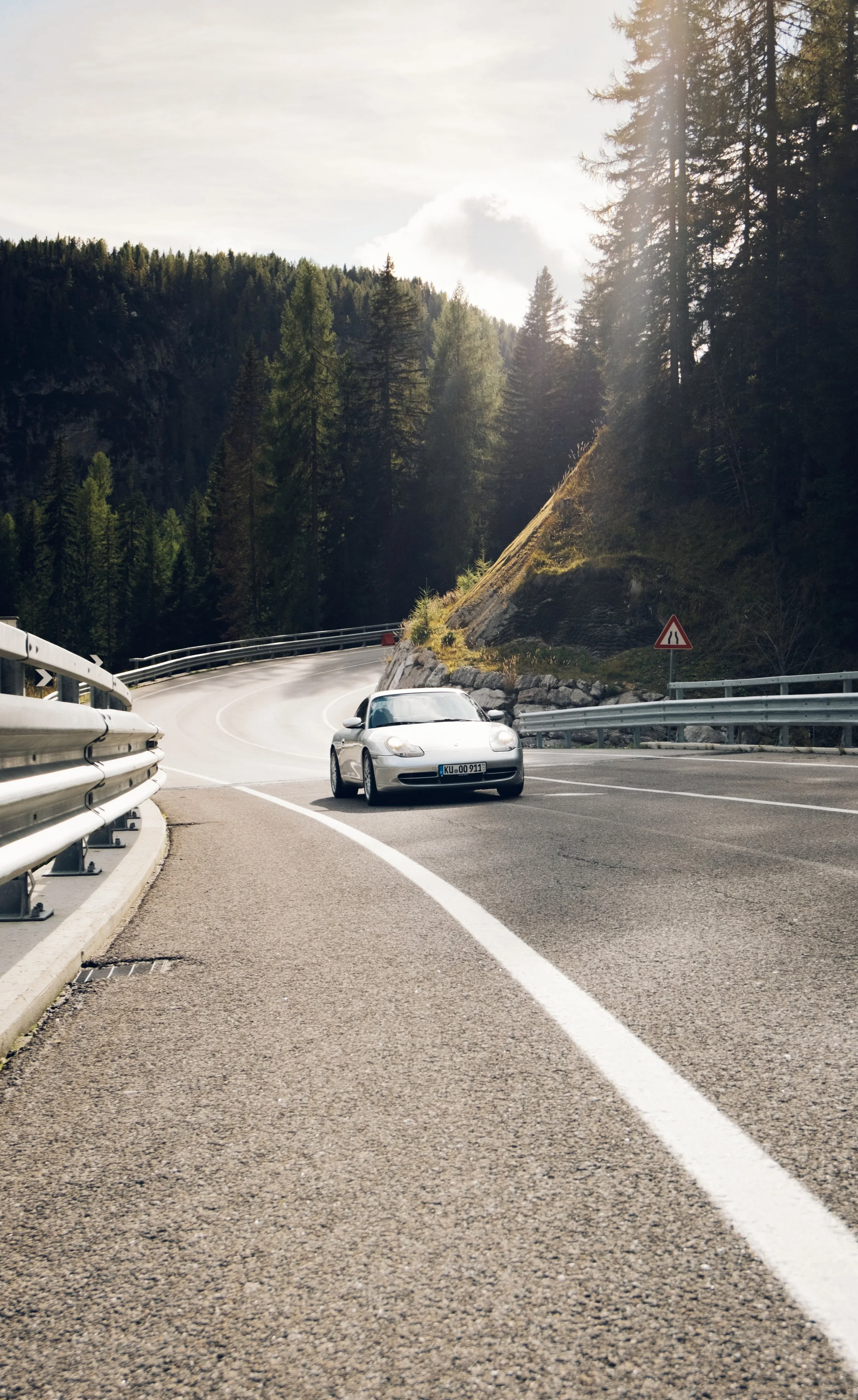 Ein silberner Sportwagen fährt eine kurvige Bergstraße mit Sicherheitsbarrieren und warnschildern, umgeben von einem dichten Wald und Hügeln unter einem bewölkten Himmel