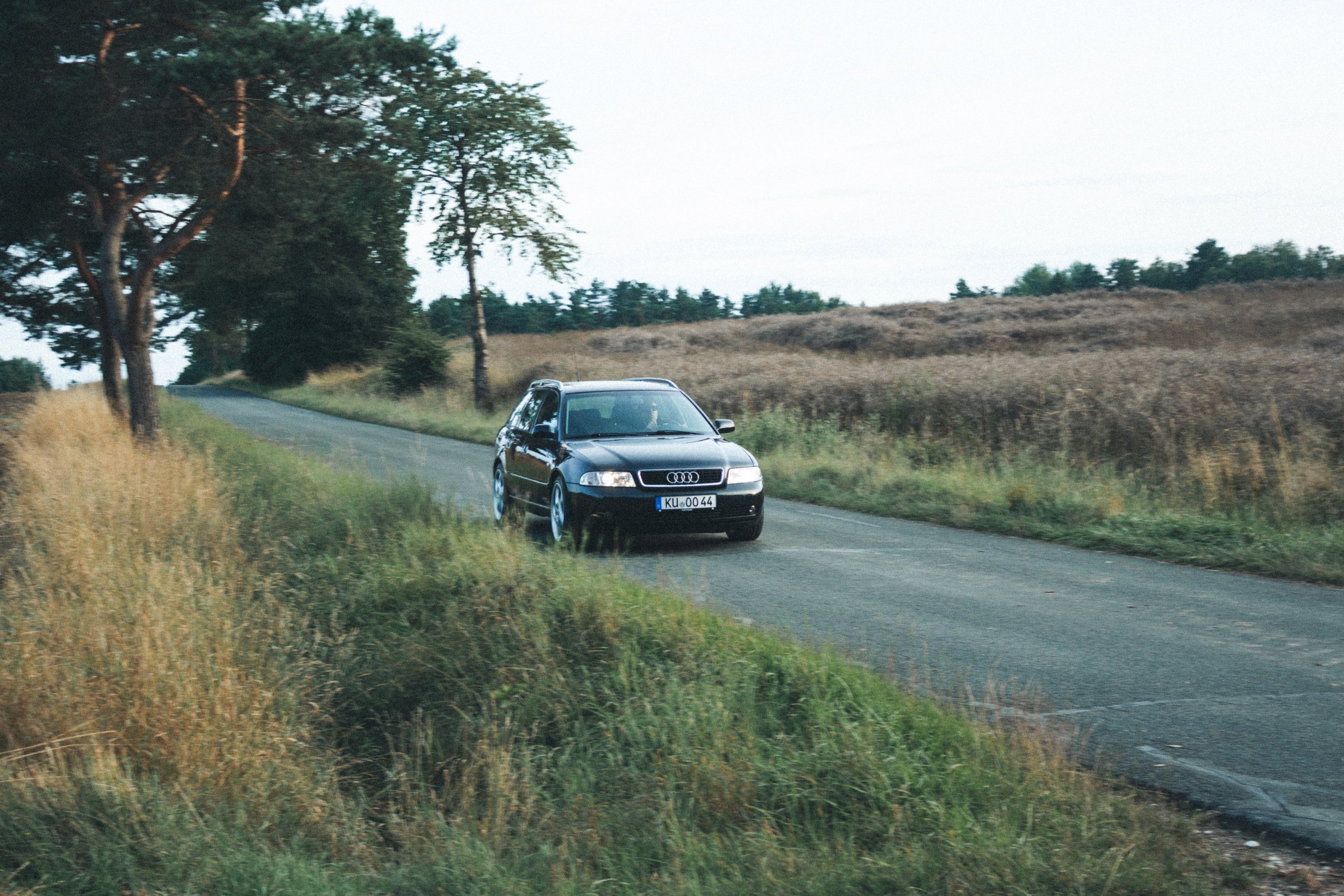 Ein schwarzer Audi-Autofährt auf einer ländlichen Straße, umgeben von grünen Feldern und Bäumen, bei bewölktem Himmel.