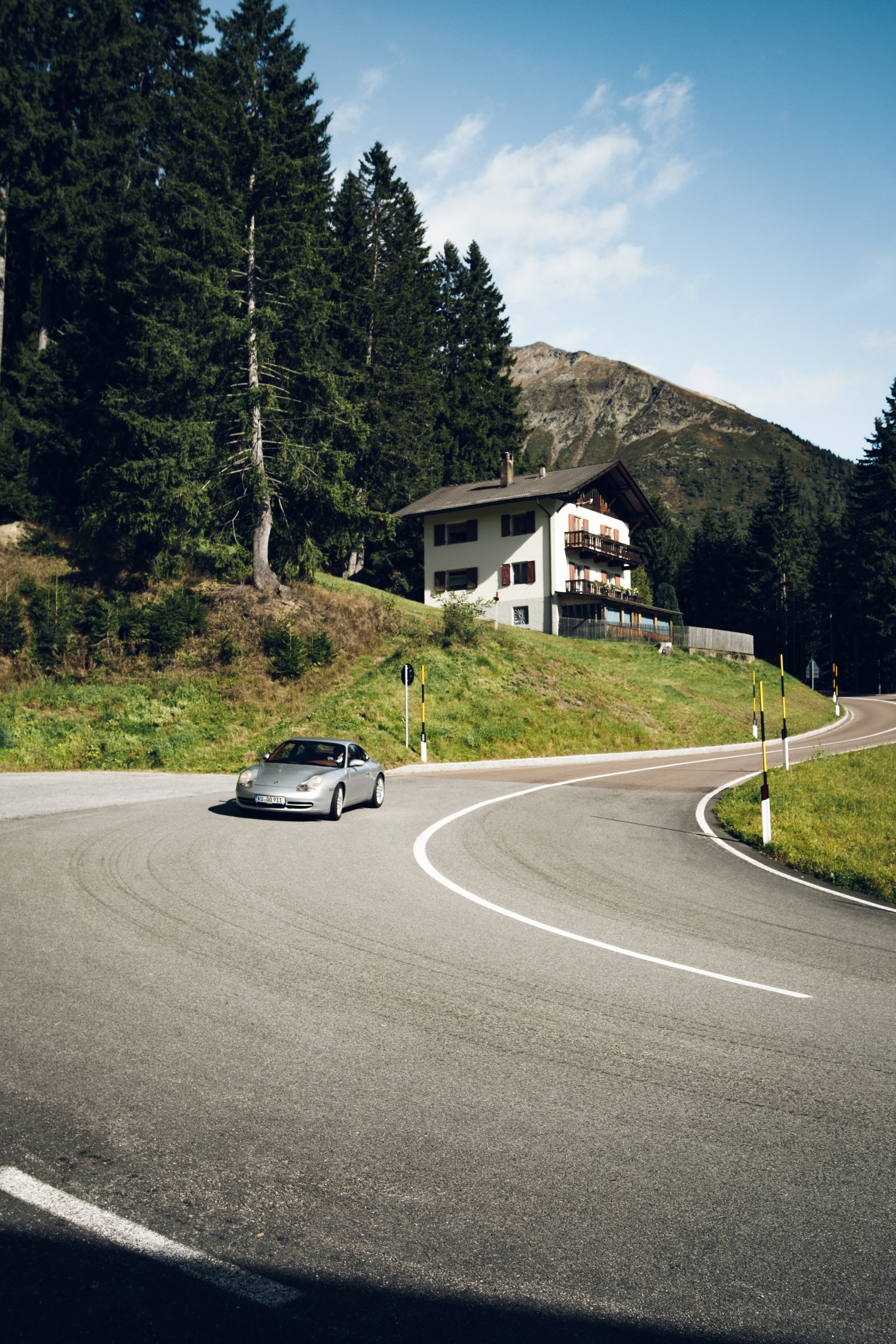 Ein kurviger Bergstraße mit einem silbernen Auto und einem Haus im Hintergrund, umgeben von Bäumen und Bergen, bei schönem Wetter.