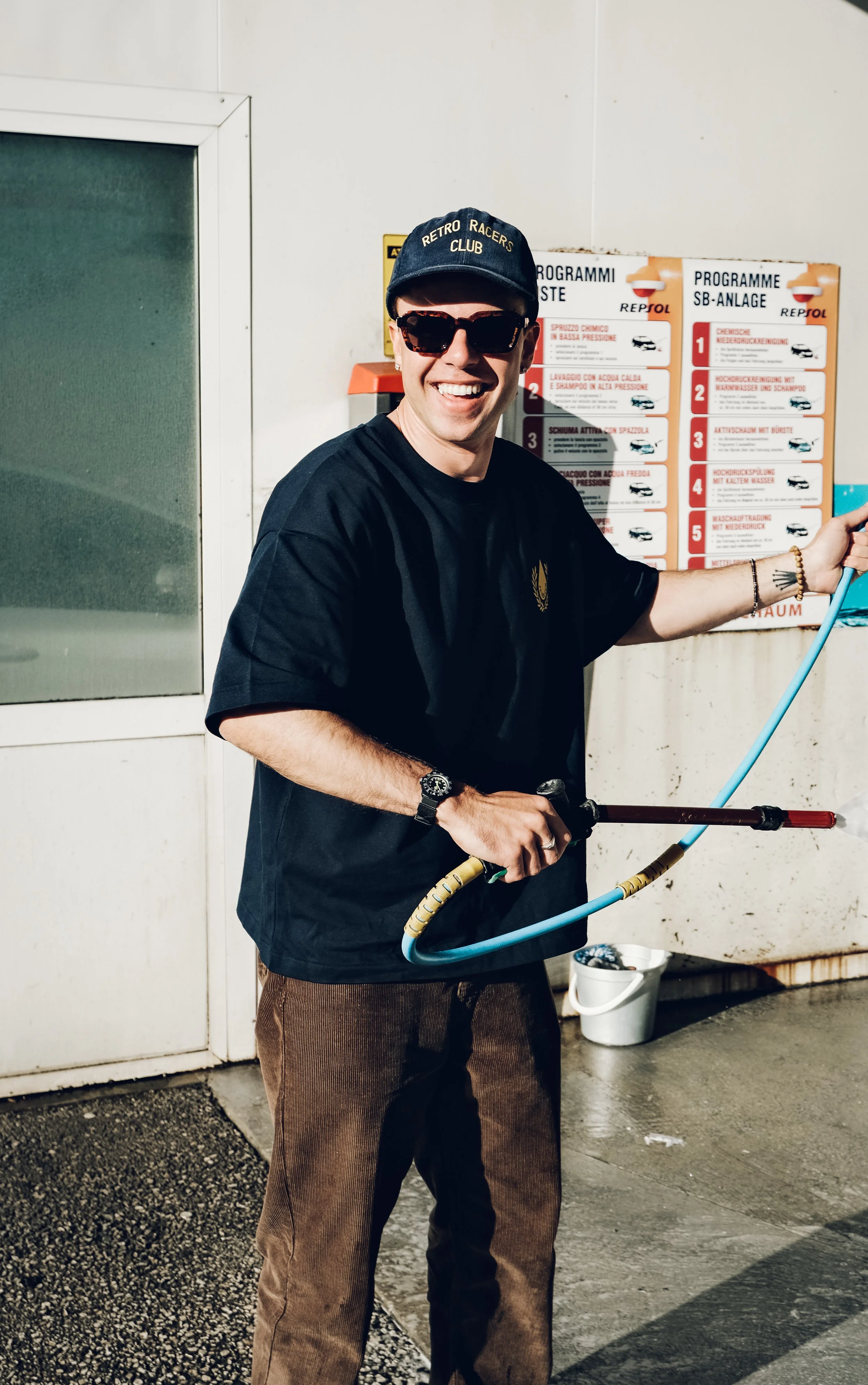 Junge Person mit Sonnenbrille und Baseballcap, die einen Hochdruckreiniger hält und bei der Autowäsche ist.