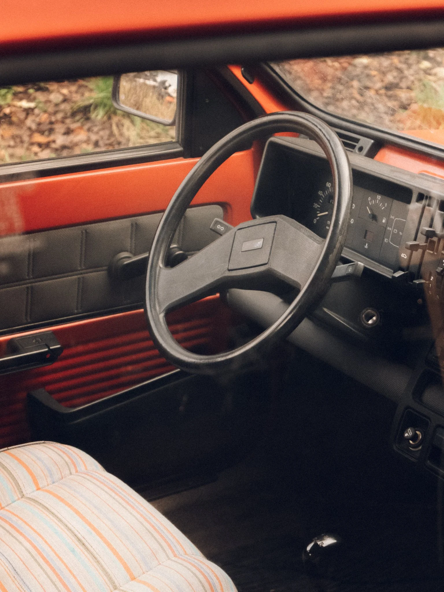 Innenraum eines alten roten Autos, Blick auf das Lenkrad und den Tachometer, mit grüner Natur im Hintergrund.