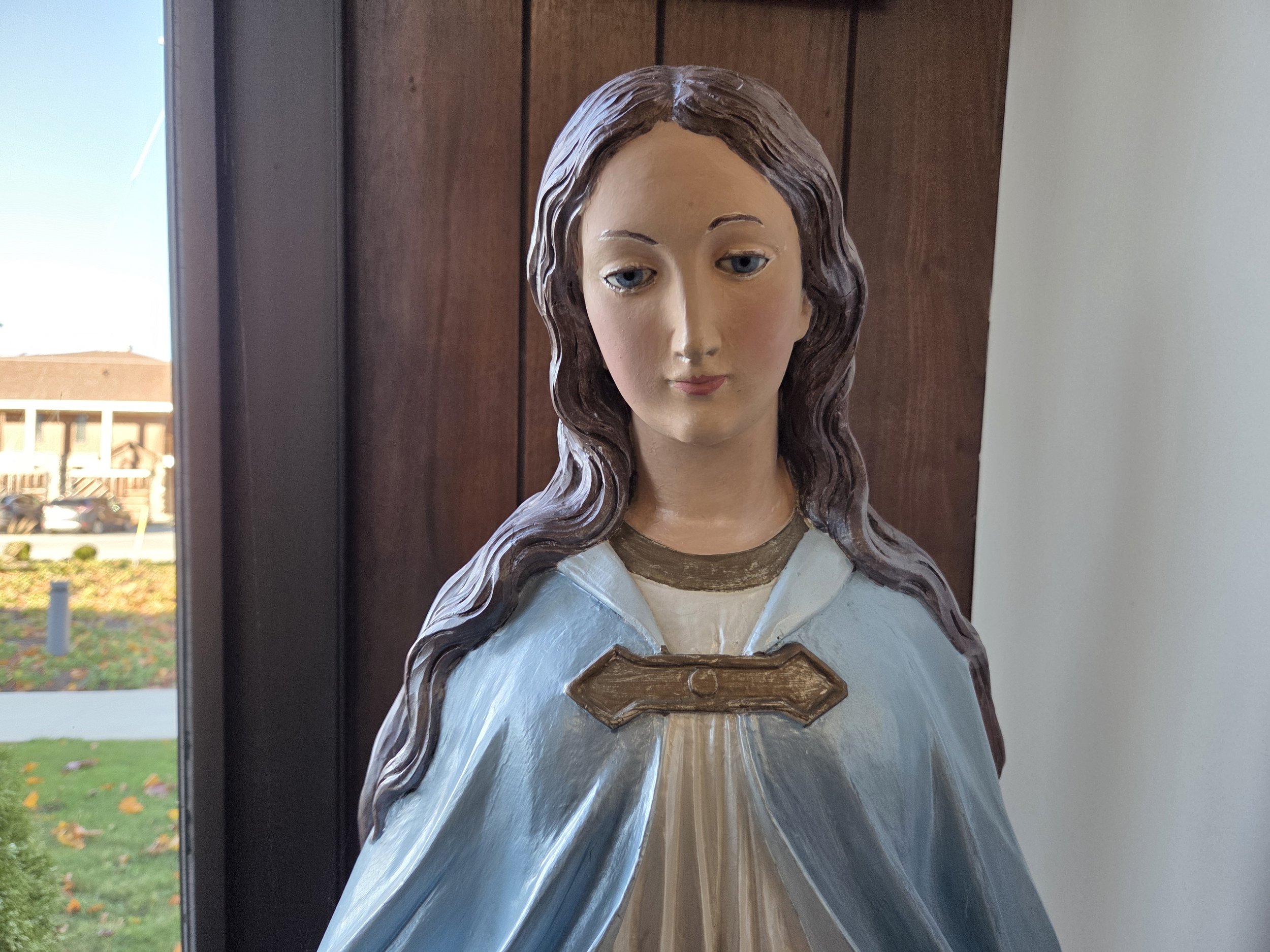 Painting of the Virgin Mary with long brown hair, wearing a light blue and beige robe, standing indoors near a window.