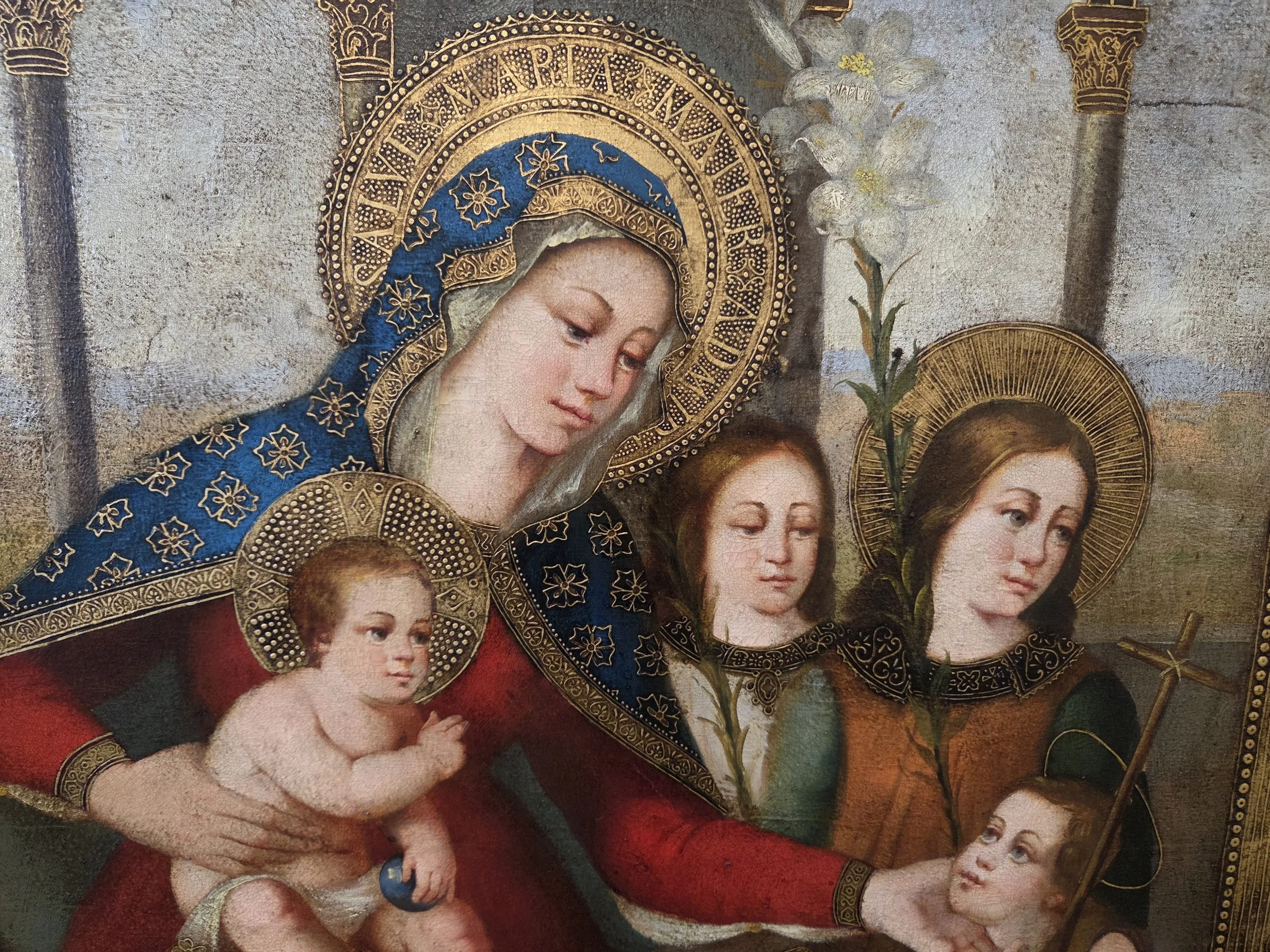 Religious painting of the Virgin Mary with baby Jesus and two young women, adorned with gold halos and intricate clothing, holding lilies and a hanging cross.