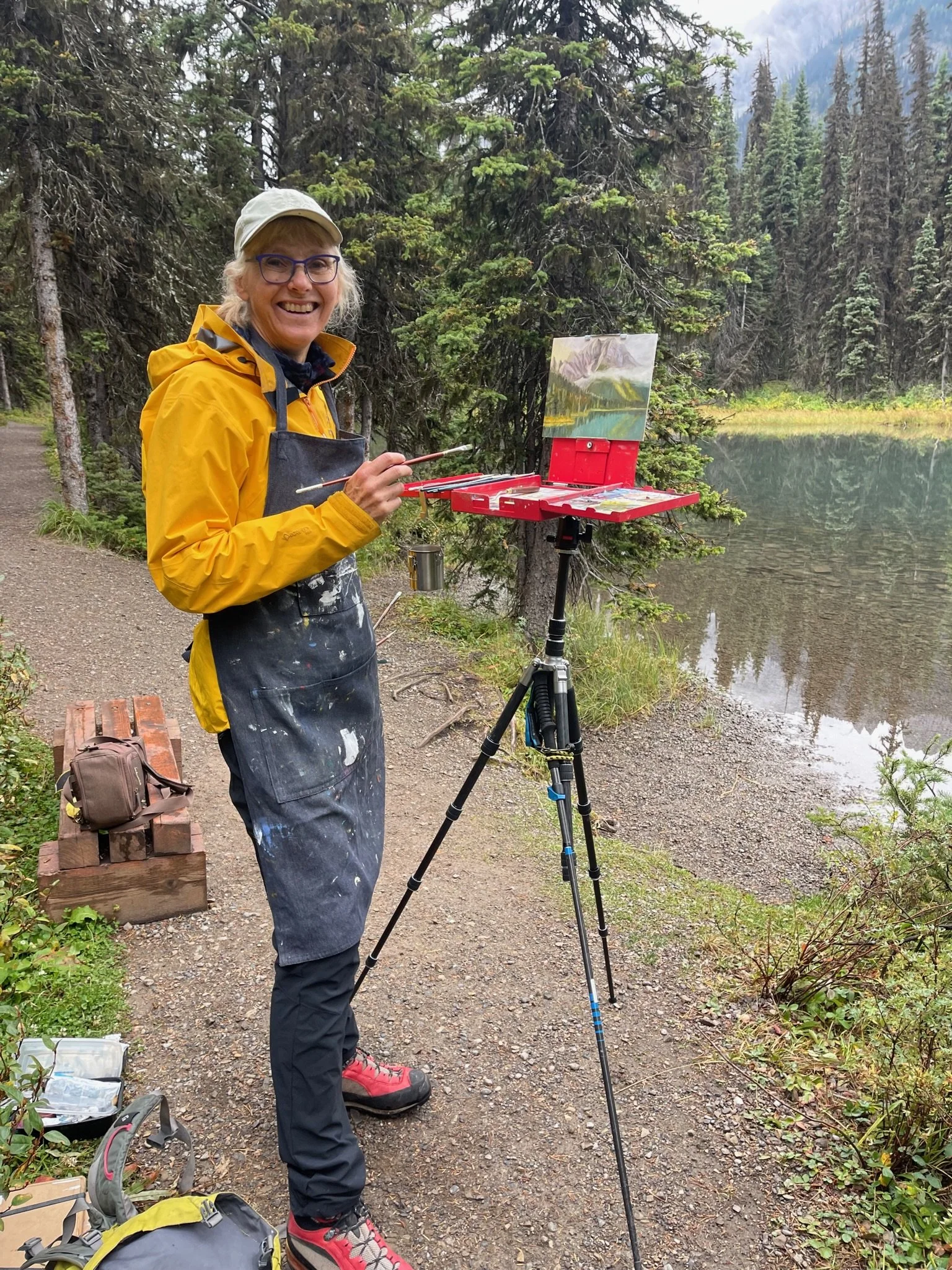 A woman in a yellow jacket and apron, standing outdoors by a lakeside with trees in the background, painting on a small canvas setup on an easel, smiling at the camera.