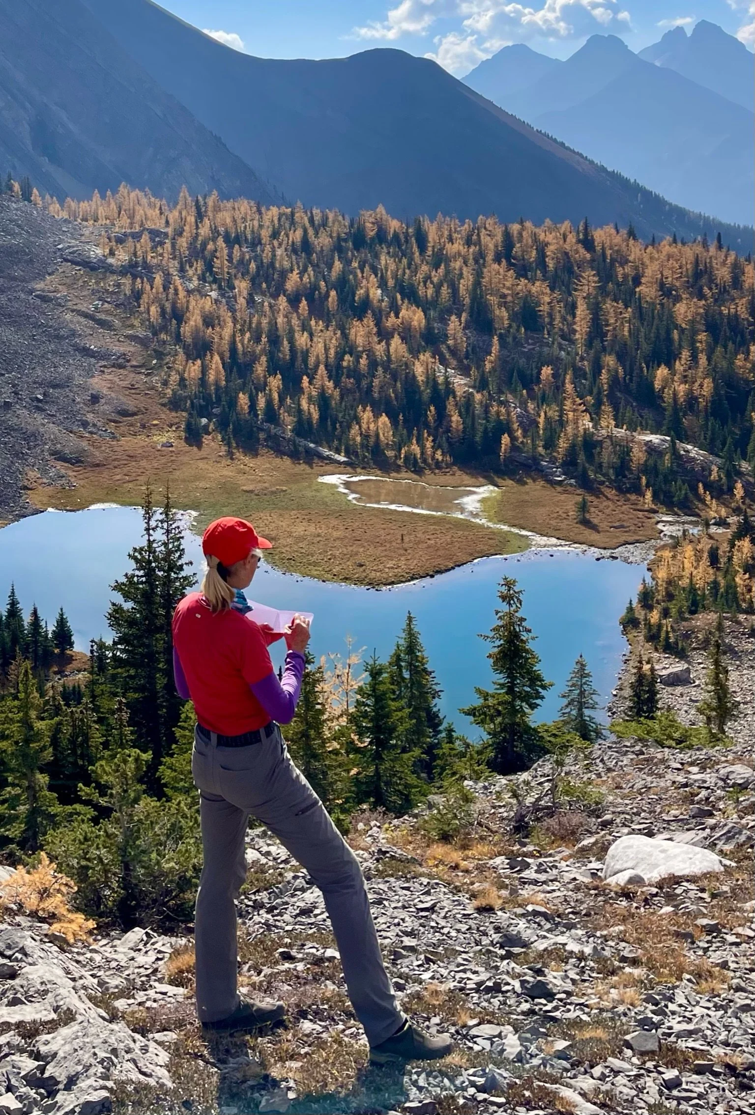 A woman in outdoor hiking clothes, wearing a red hat and purple undershirt, stands on rocky ground overlooking a mountain lake surrounded by trees and mountains, taking notes or drawing in a notepad.
