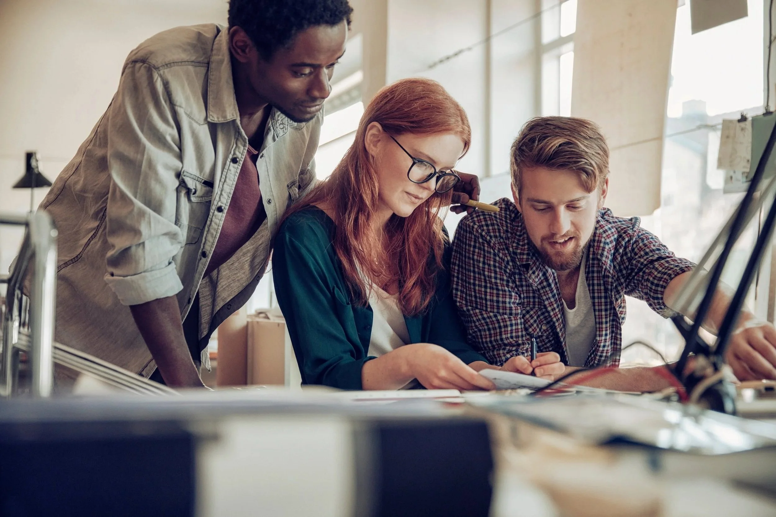 Three young adults collaborating at a table in a bright, modern workspace, with one woman writing and two men leaning in to observe.