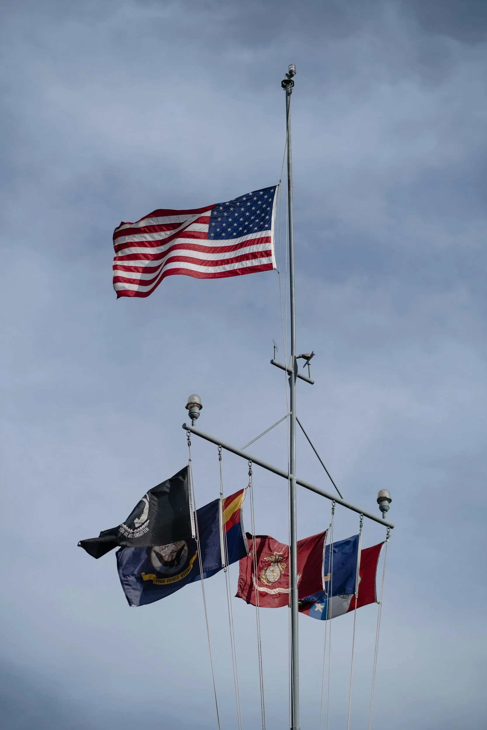 American flag flying at the top of flagpole with multiple other flags, including military and state flags, hanging below against cloudy sky.