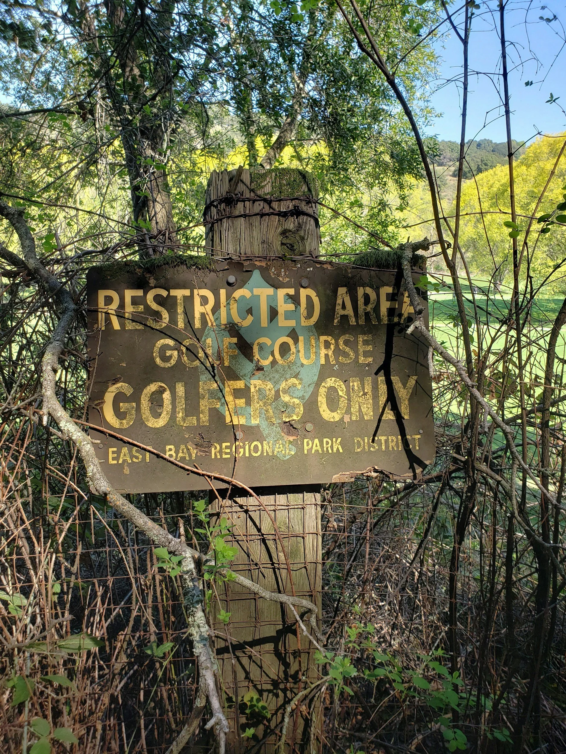 A weathered sign surrounded by overgrown vegetation, indicating a restricted golf course for golfers only, located in East Bay Regional Park District.