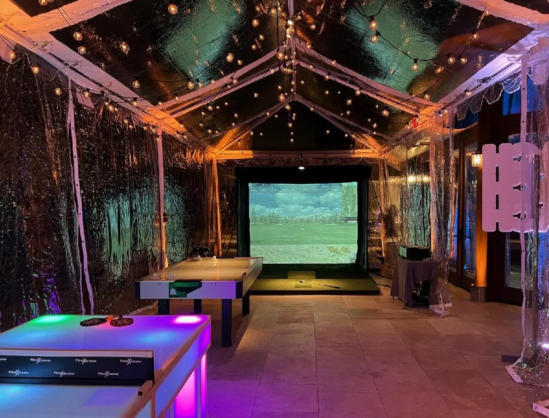 Indoor event space decorated with string lights, shiny metallic walls, a projector screen showing an outdoor field scene, and various gaming tables with colorful lighting.