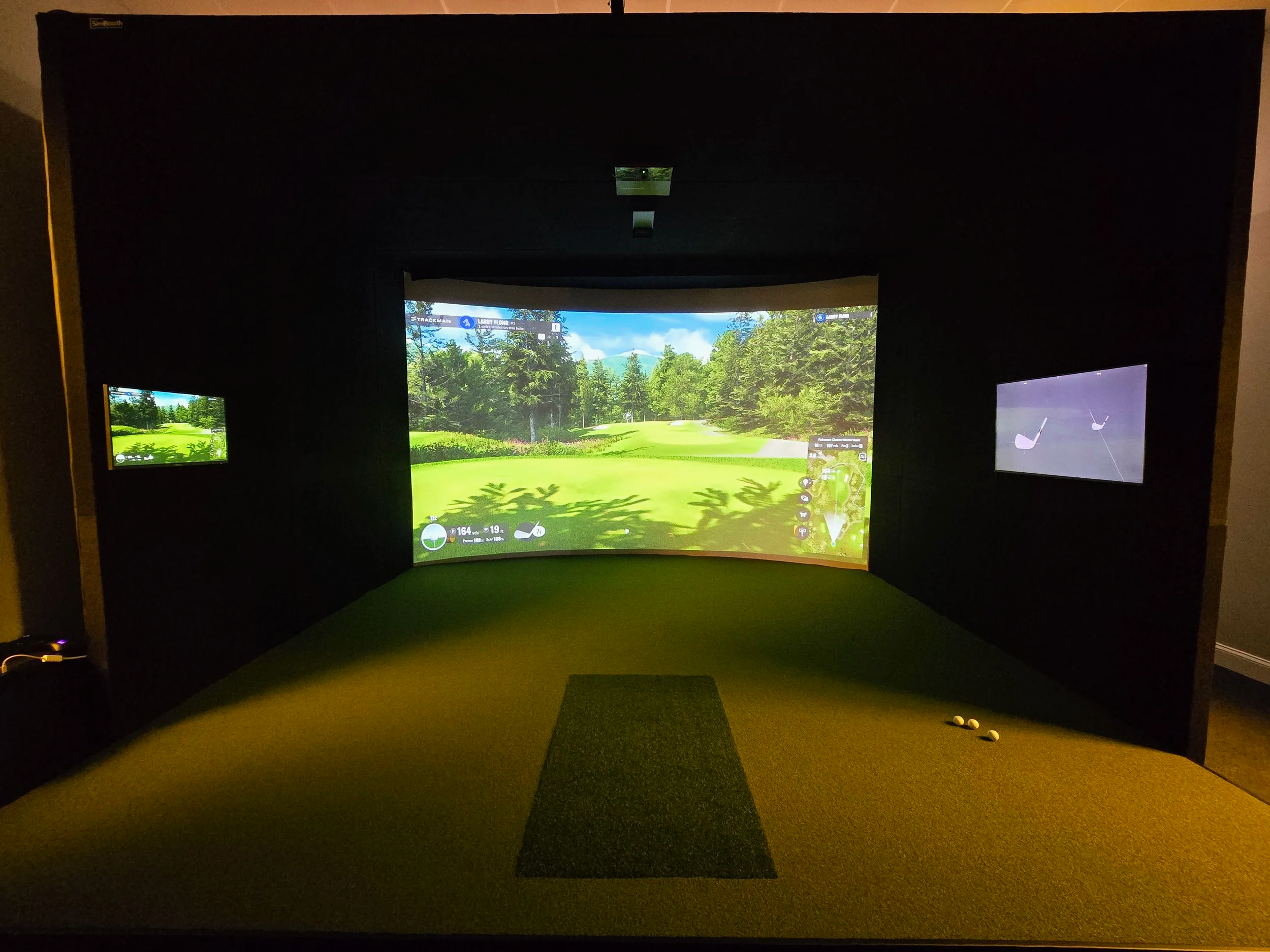 Indoor golf simulation room with a large curved screen displaying a golf course, two smaller monitors on the sides, and some golf balls on the floor.