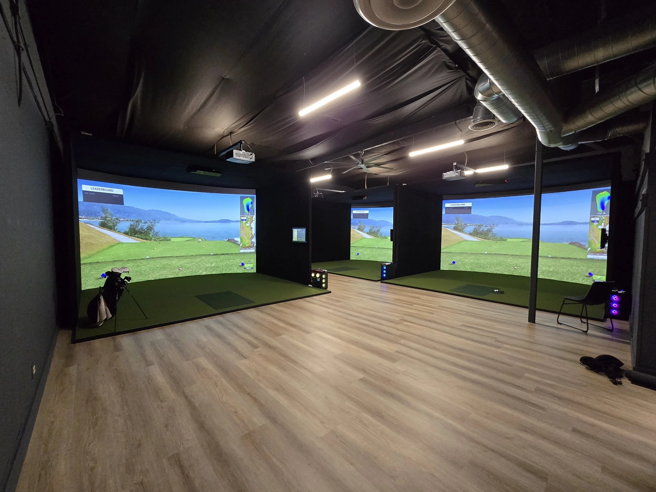 Indoor golf simulation room with three screens showing a virtual golf course. Each station has a putting green, golf clubs, and seating area.