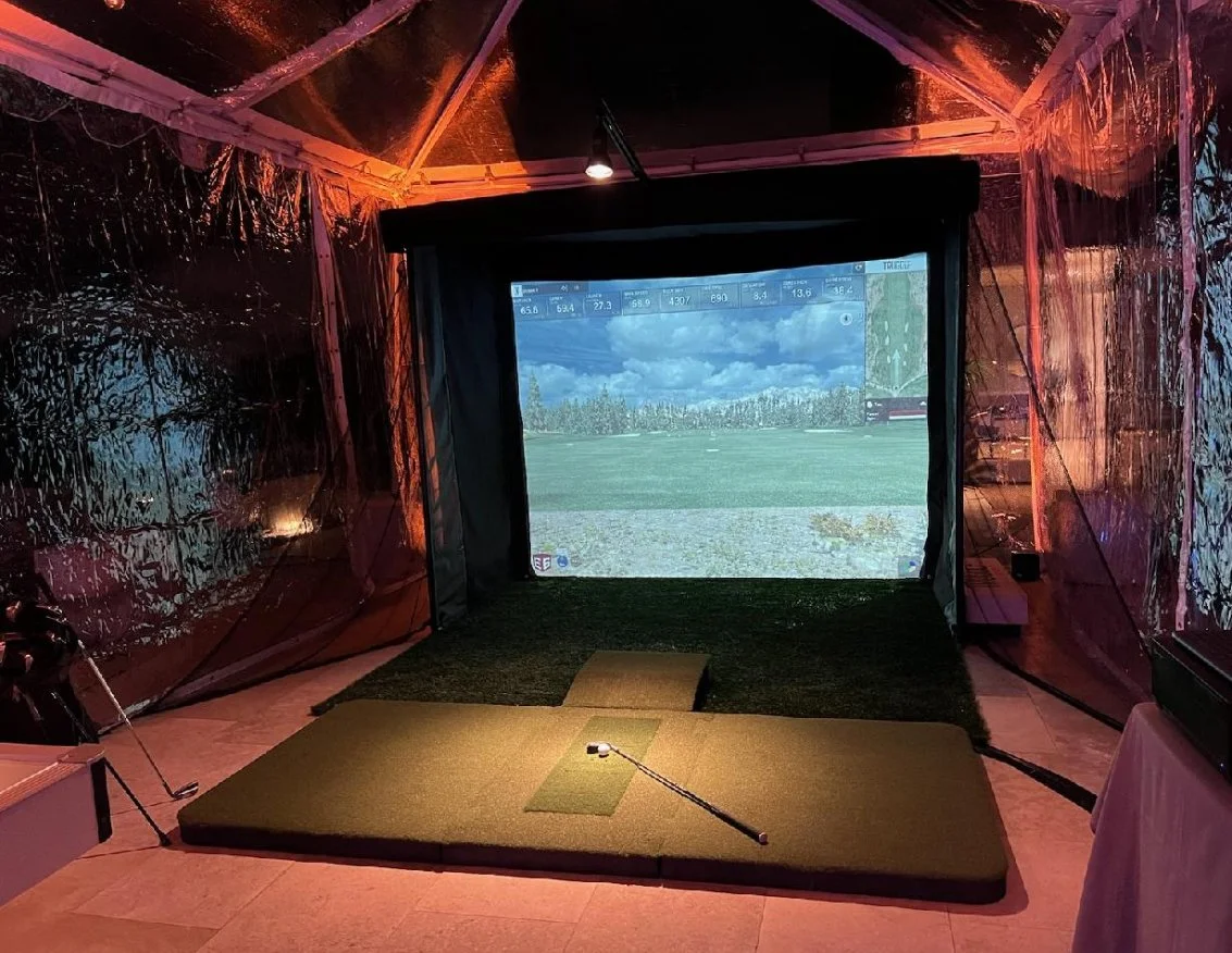 Indoor golf simulator with a giant screen displaying a golf course and a golf club on a padded mat.