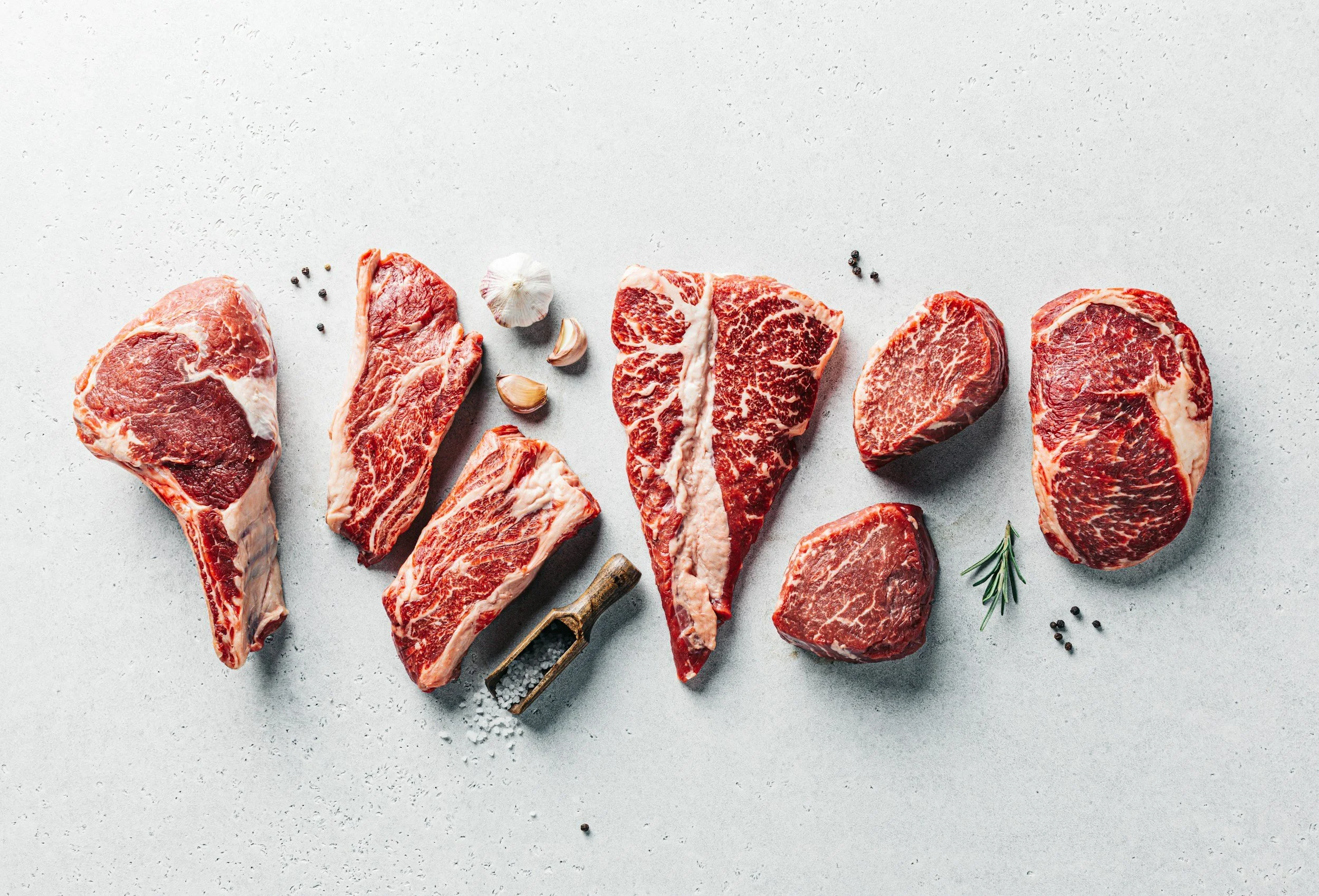 Various cuts of raw beef steak arranged on a light gray surface, garnished with garlic cloves, rosemary, black peppercorns, and sea salt.