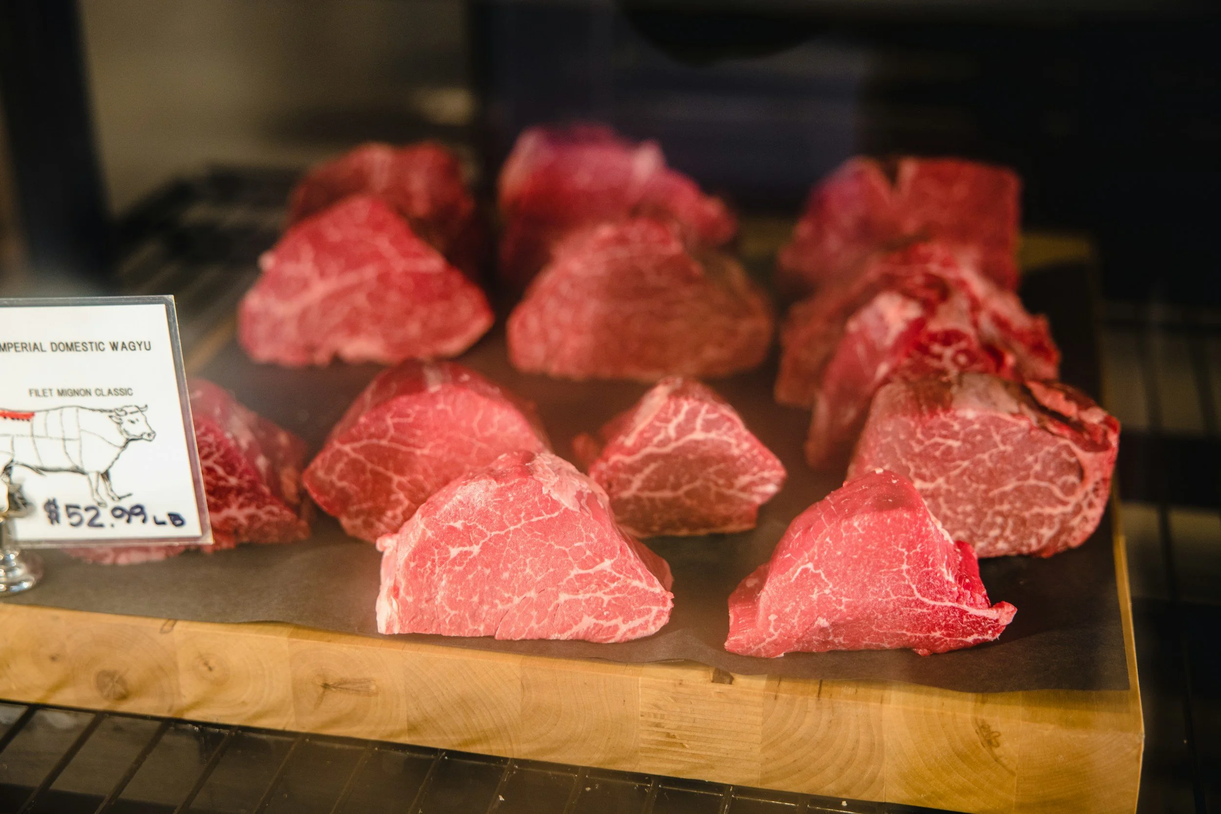 Several pieces of marbled raw beef on a wooden cutting board at a butcher shop, with a sign indicating the meat is Imperial Domestic Wagyu, priced at $52.99 per pound.