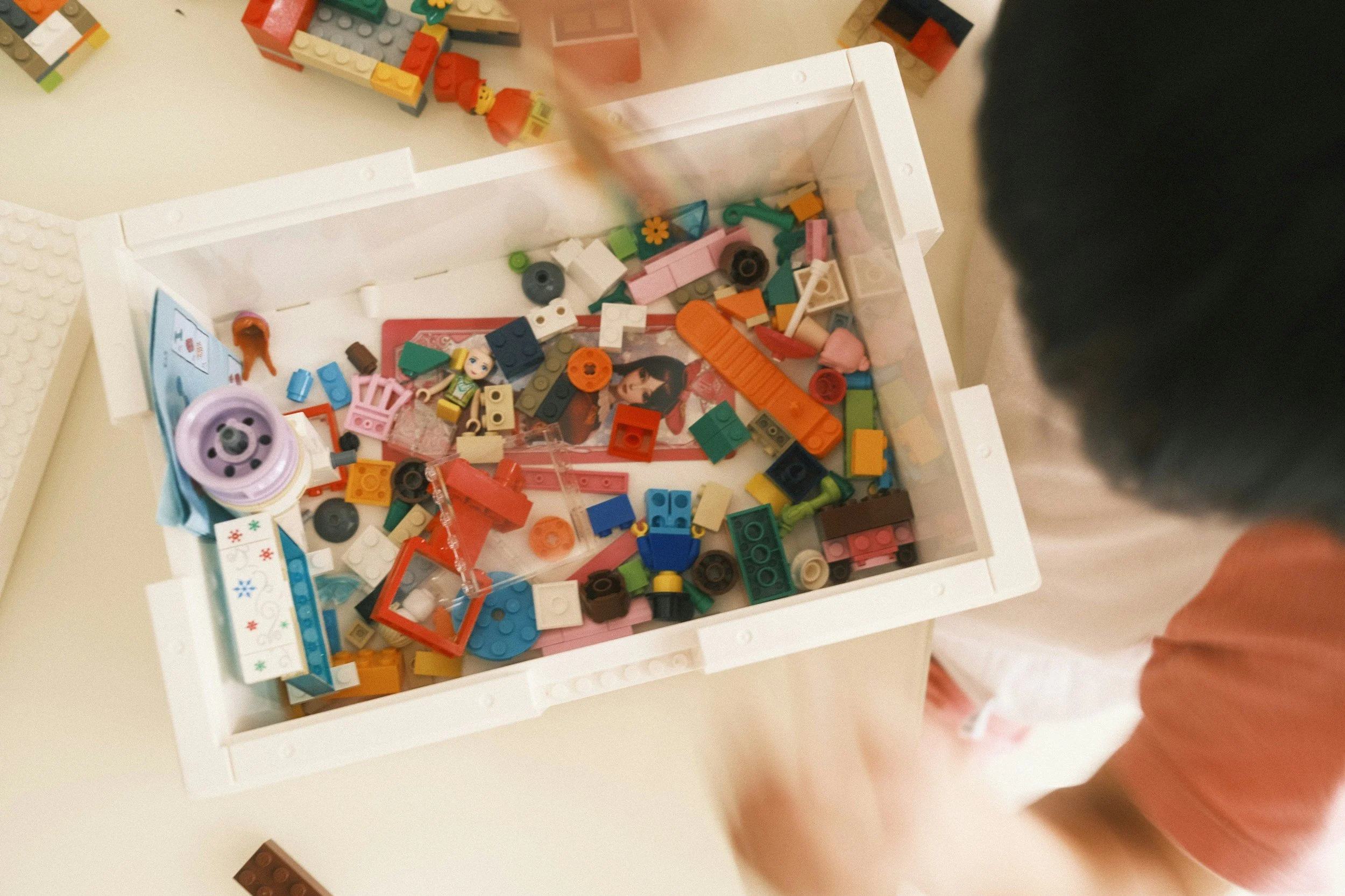 Child playing with assorted LEGO bricks and toys in a white storage box on a table.