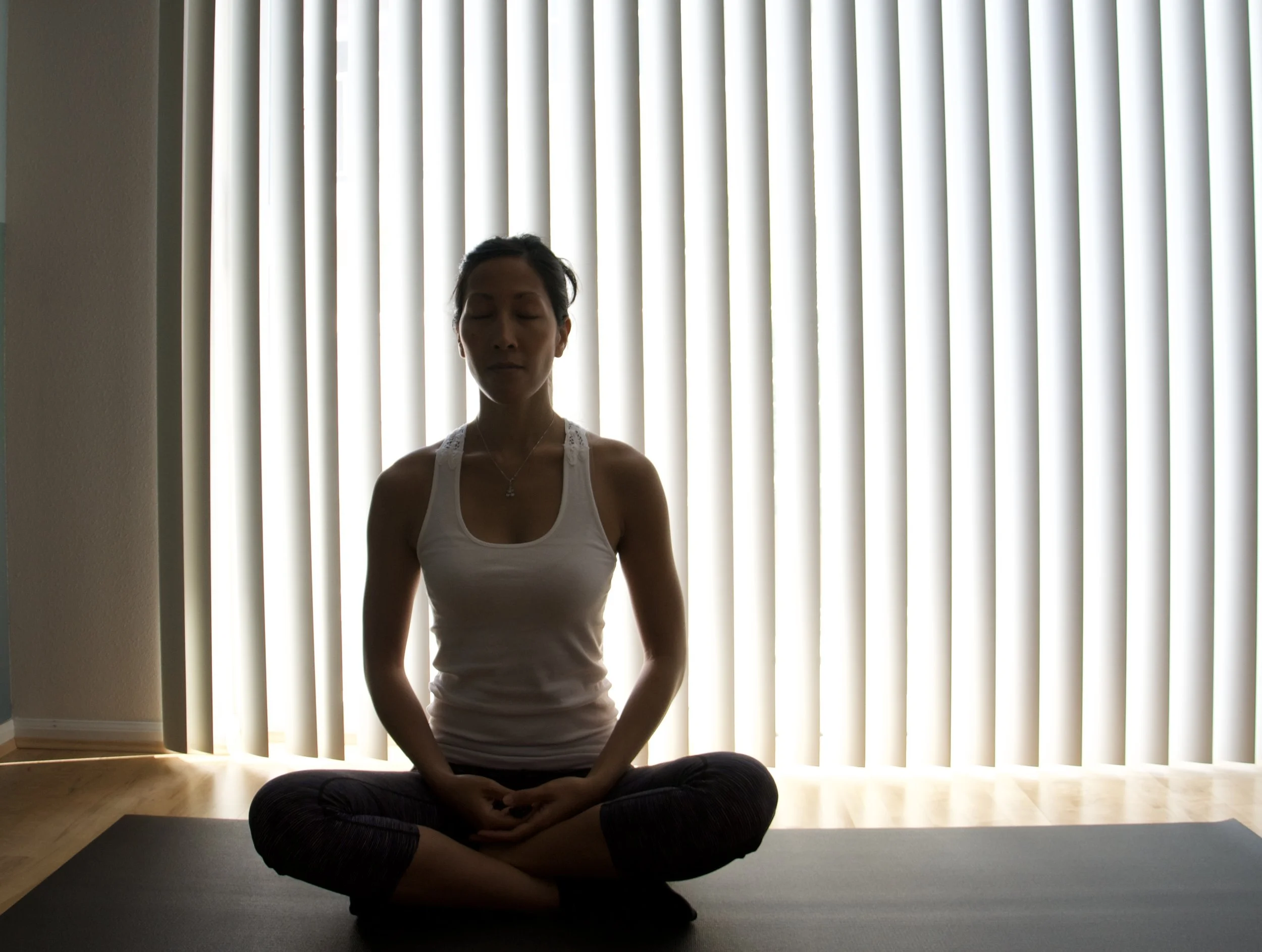 A woman sitting cross-legged on a yoga mat in front of vertical blinds, meditating with eyes closed.