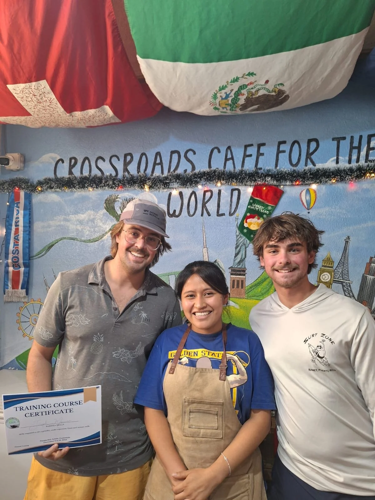 Three smiling people standing in front of a colorful mural that reads 'Crossroads Cafe for the World'. The person on the left is holding a certificate, and the person in the middle is wearing an apron.