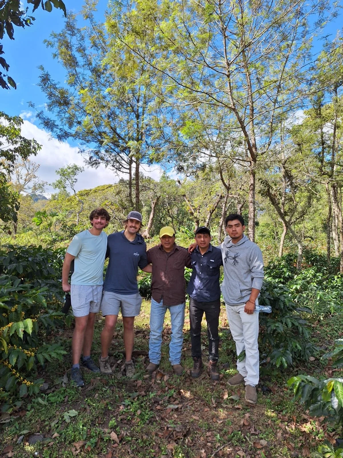 Five men standing closely together outdoors in a forested area with trees and leafy greenery, smiling at the camera.