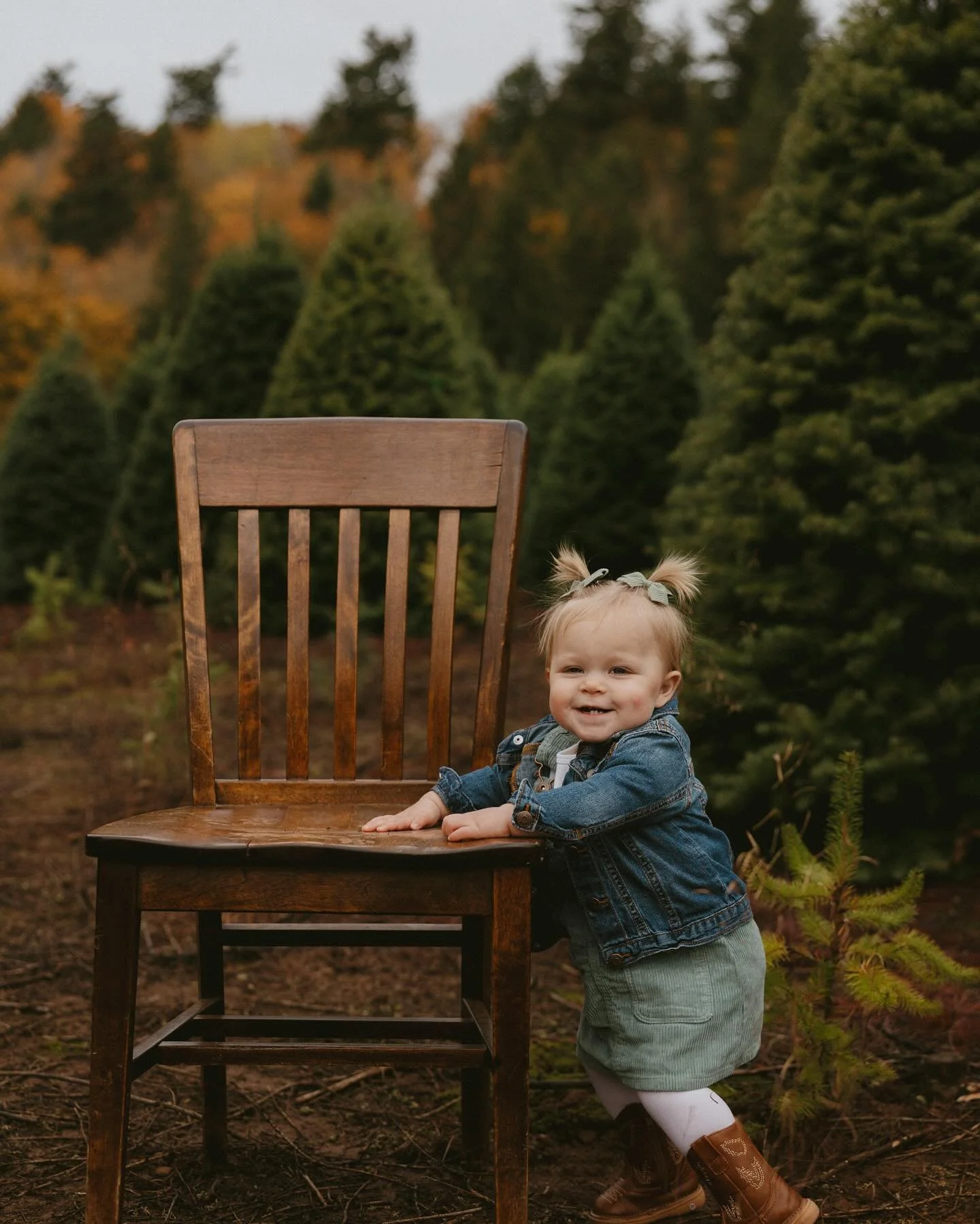 A young girl standing on the ground outside, leaning on a wooden chair, smiling, surrounded by evergreen trees and fall foliage.