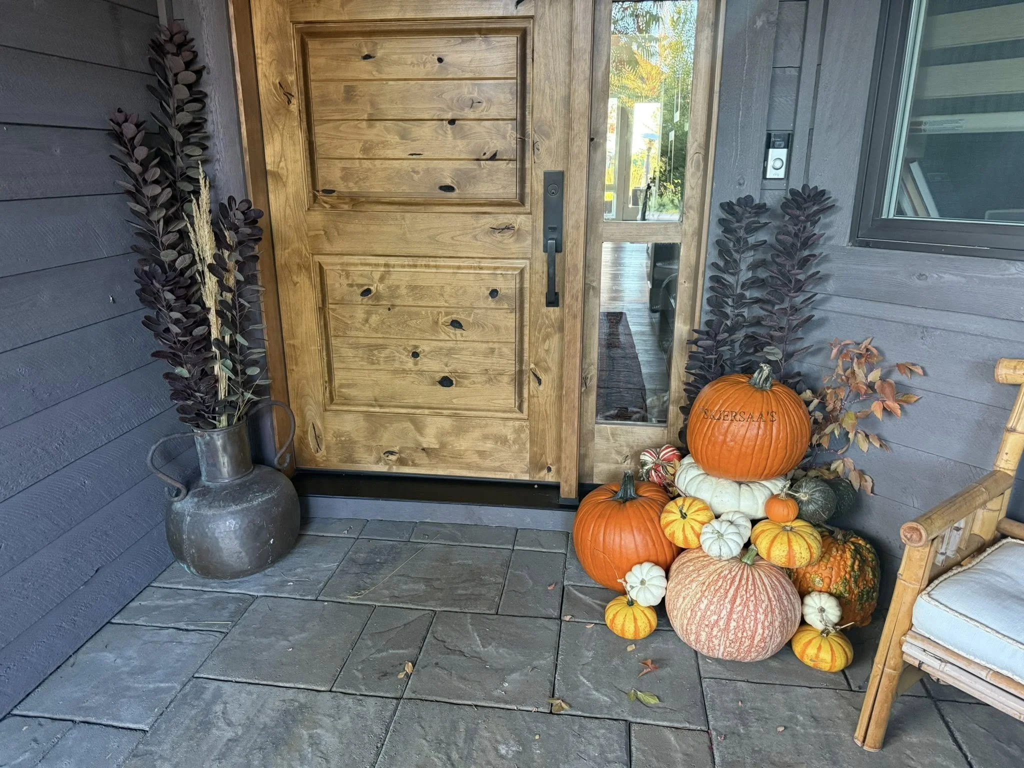 Porch entryway with a wooden door, potted dark purple leaves on both sides, and a display of various pumpkins in different sizes and colors, including orange, white, and green, with some small striped ones, arranged on the porch corner. A vintage met
