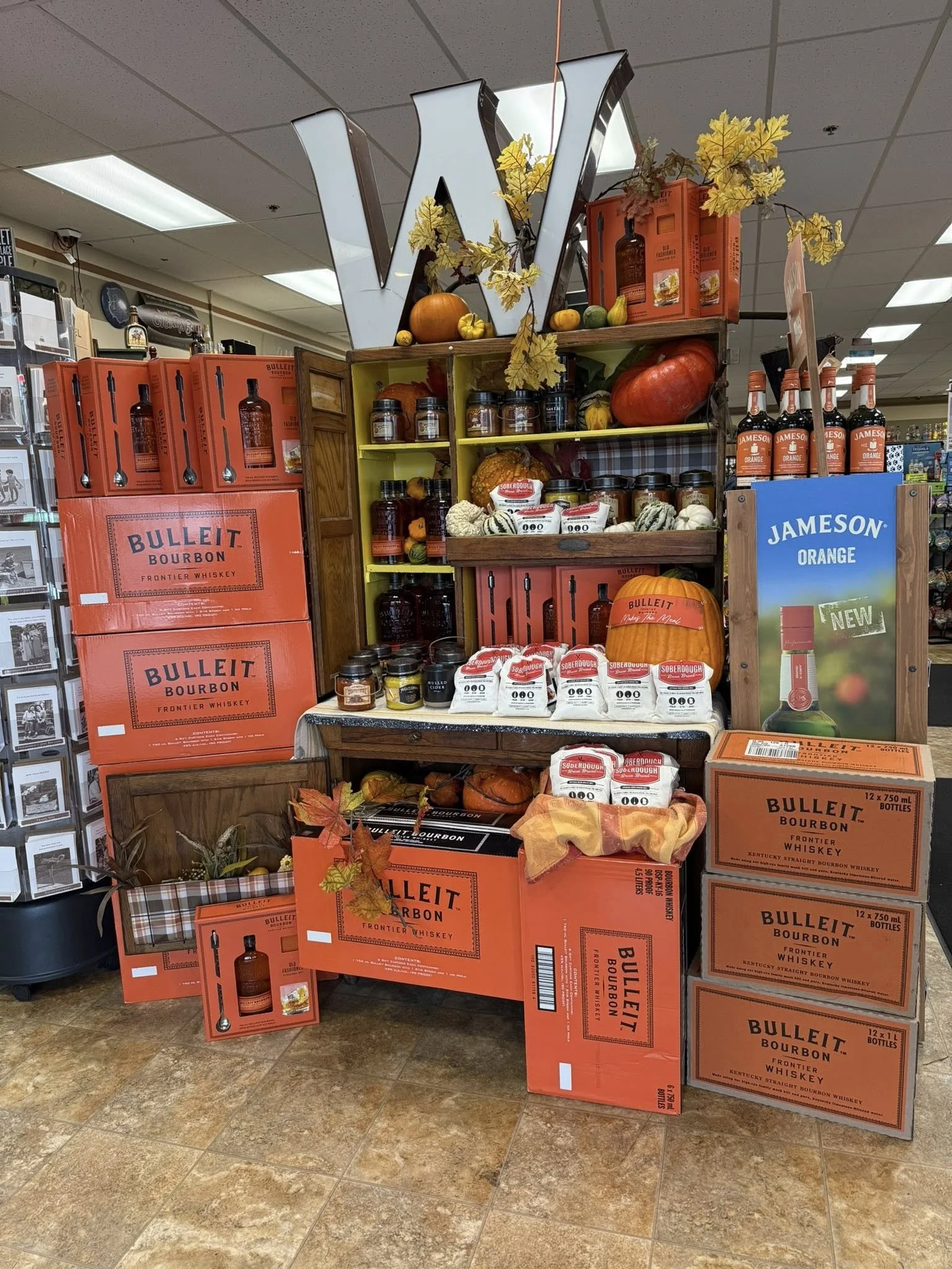 Display of Bulleit bourbon whiskey boxes with autumnal decorations, pumpkins, gourds, and jars, set up on a store shelf in an indoor retail space.