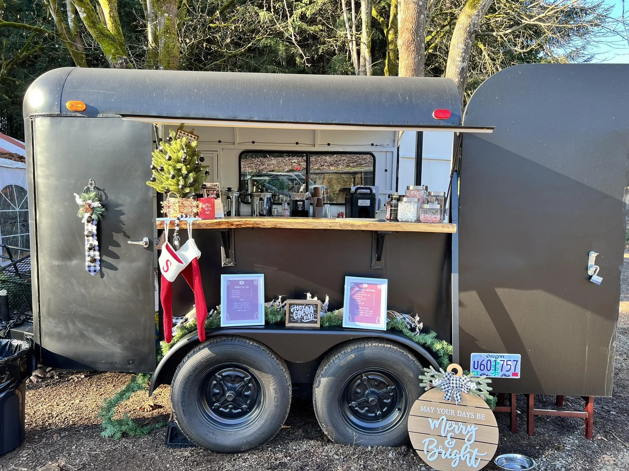 A black food trailer decorated for Christmas, featuring holiday ornaments, a small Christmas tree, and a wooden sign with holiday greetings. The trailer has a serving window with frames displaying menus, and various jars and containers on the counter