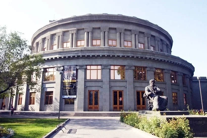 A large, round, classical-style building made of gray stone with several windows and decorative columns. A statue of a seated person is situated in front of the building, surrounded by a landscaped area with bushes and flowers. The sky is clear with some sunlight.