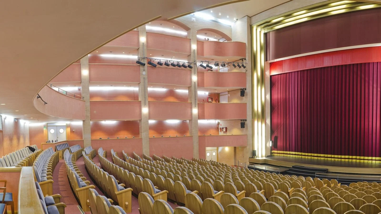 Inside a theater with rows of empty beige seats facing a large stage with a closed red curtain.