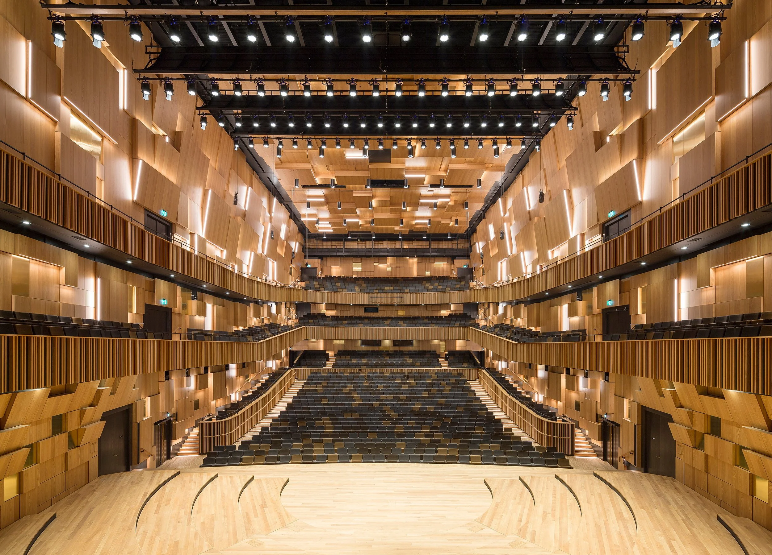 A modern concert hall with wooden paneling, tiered seating, and stage, illuminated by numerous ceiling lights.
