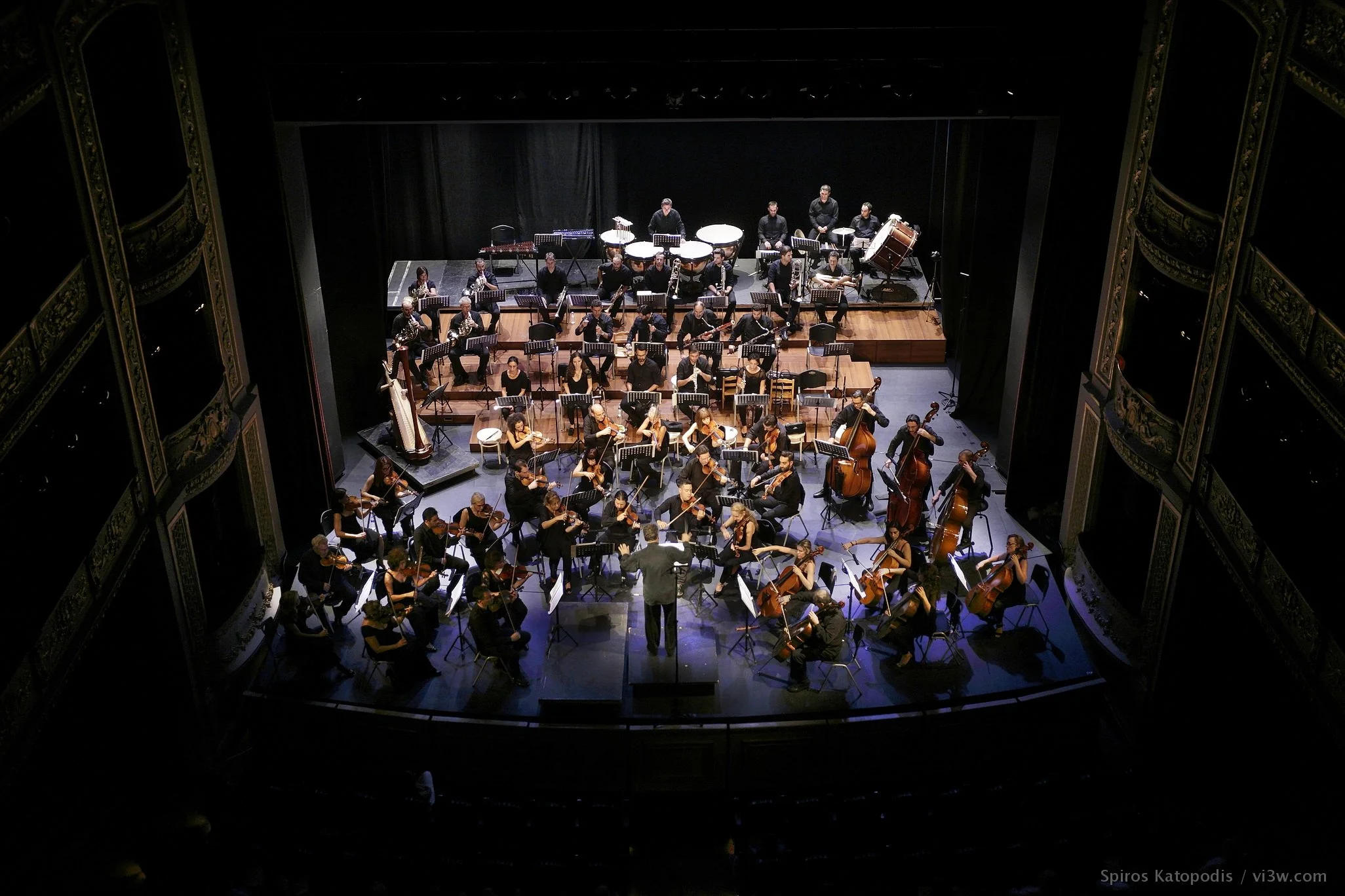 An orchestra performing on a stage in a concert hall, with musicians playing string, wind, and percussion instruments, directed by a conductor.