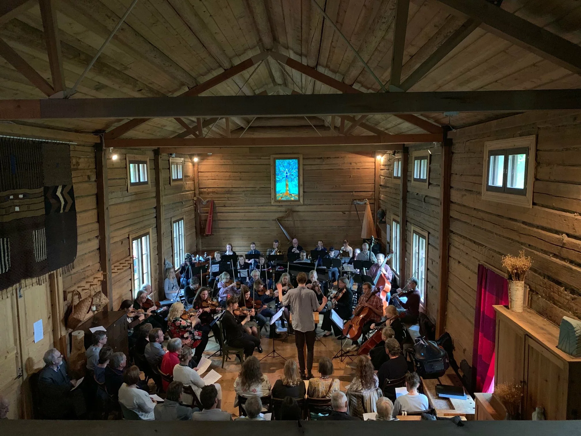 An orchestra performing in a wooden hall with audience seated around.