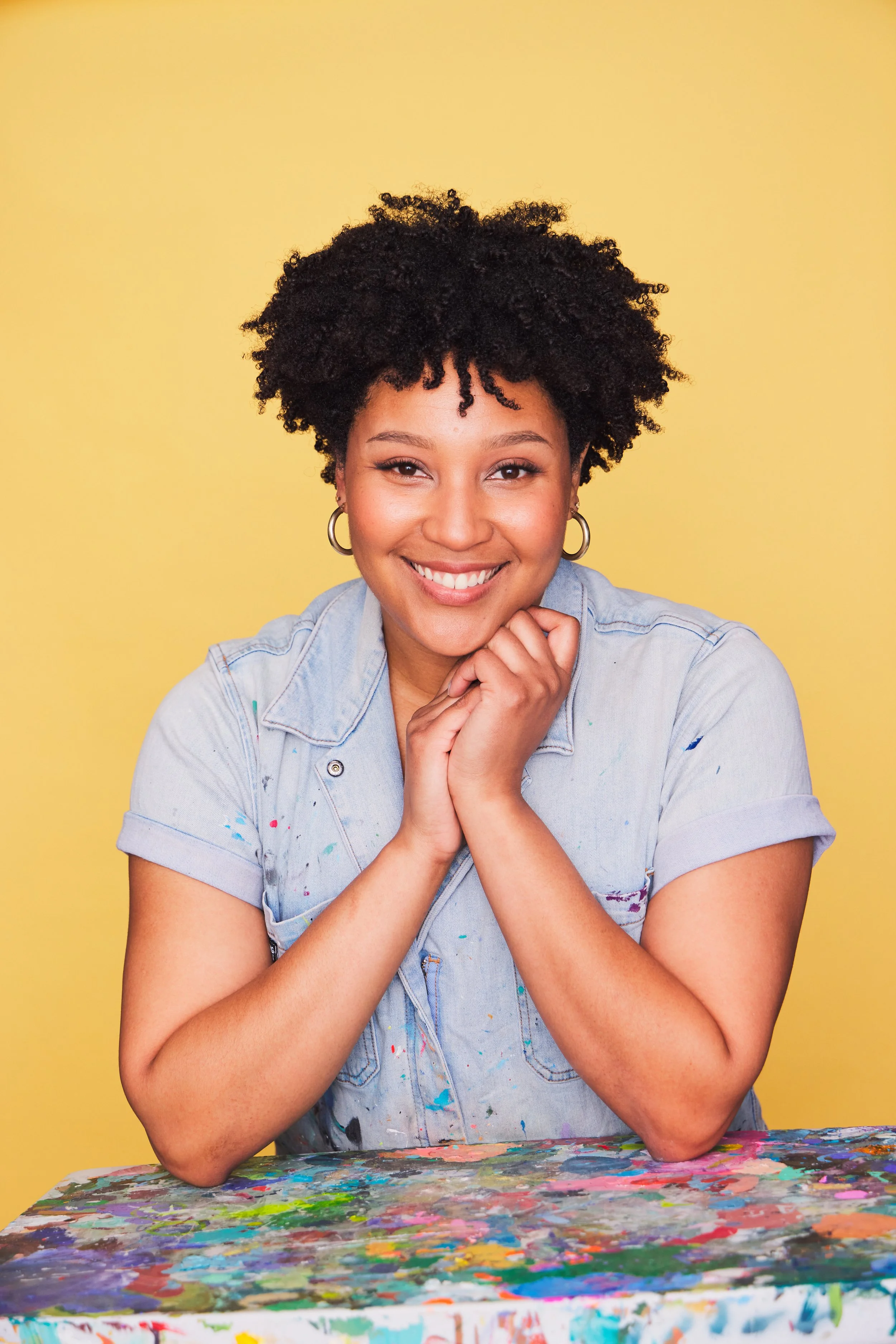 Smiling woman with natural curly hair, wearing gold hoop earrings and a light blue denim shirt, sitting at a table with colorful paint splatters, against a yellow background.