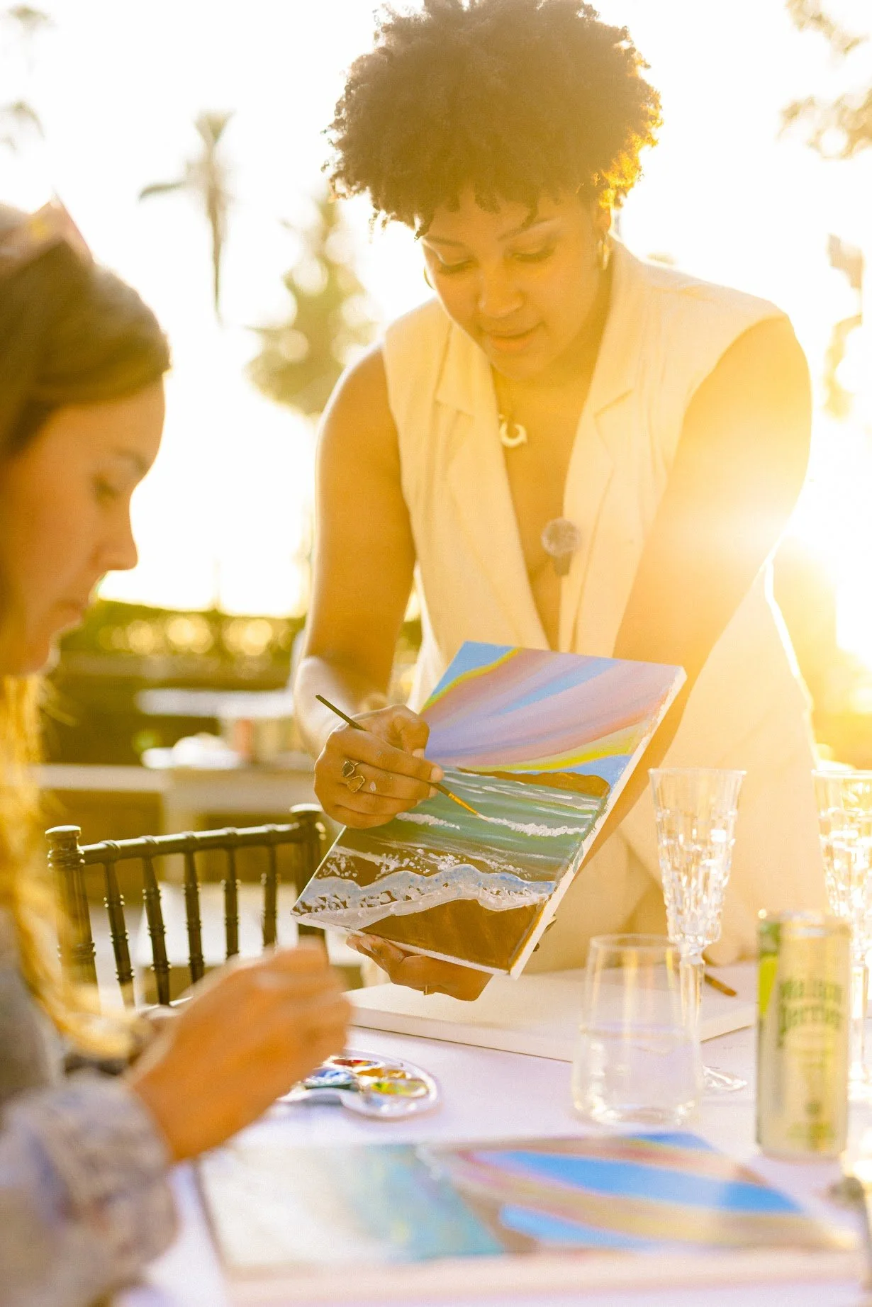 Woman in a sleeveless white top painting a seascape on a canvas at an outdoor event, with another person partially seen in the foreground. The scene is backlit by bright sunlight, creating a warm glow.