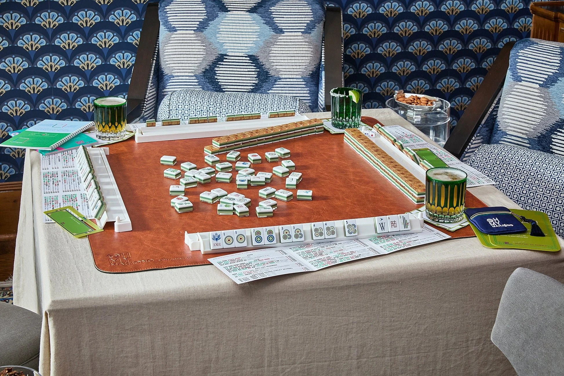 An active Mahjong game set up on a dining table with game tiles, drinks, and score sheets surrounded by cushioned chairs with patterned upholstery.