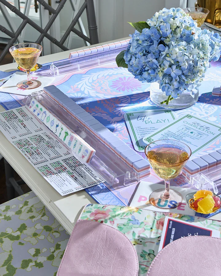 A table set for a game night with a large bouquet of blue hydrangeas, two glasses of white wine, a card game, a menu, and colorful decor in a bright, airy room.
