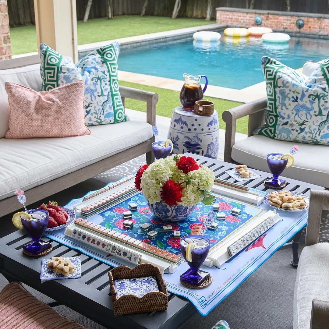 Patio with table set for a game night, featuring a colorful board game, snacks, drinks, and a bouquet of flowers, overlooking a backyard pool with floaties.
