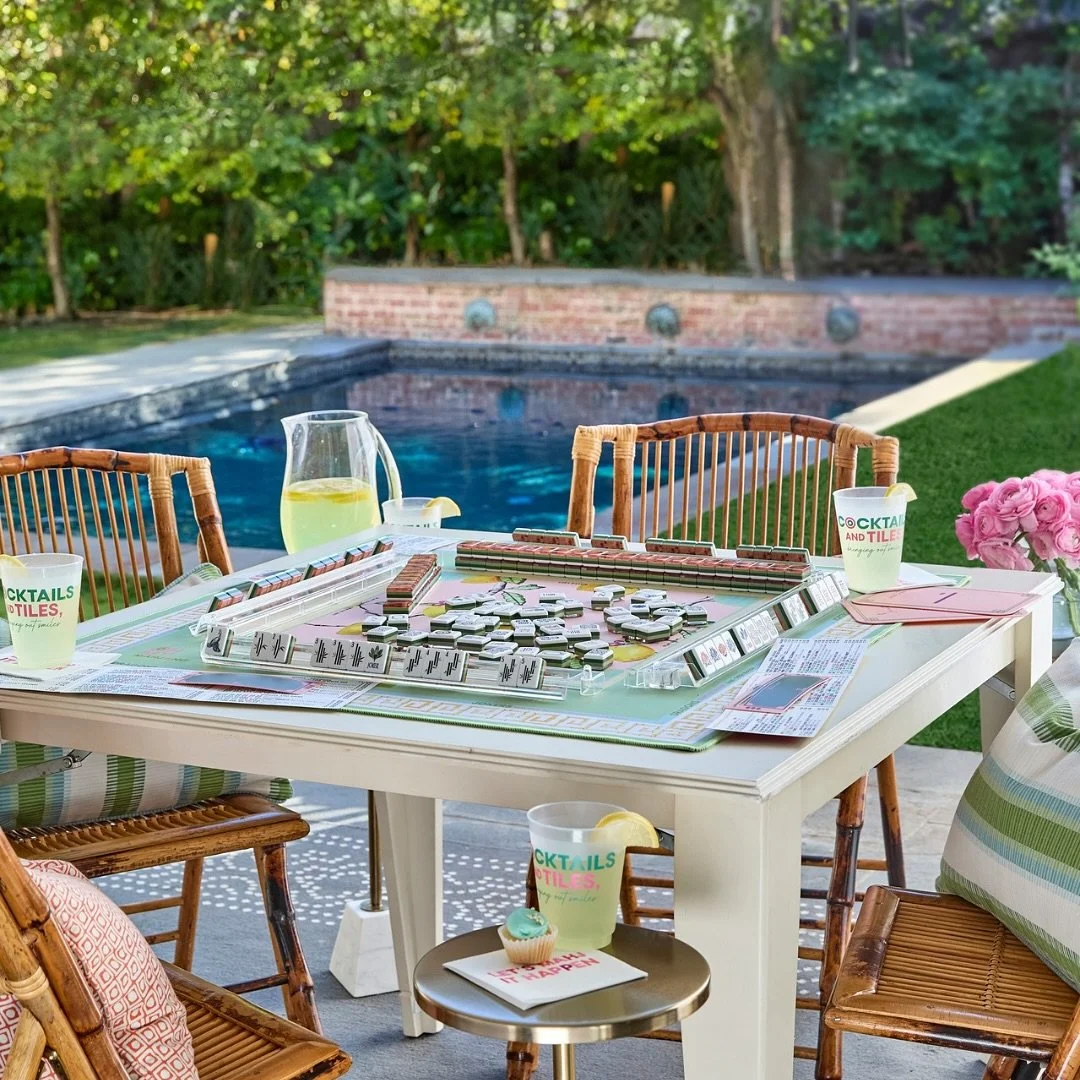 A backyard patio with a table set up for playing Monopoly, with drinks, cupcakes, and pink flowers, overlooking a swimming pool and greenery.