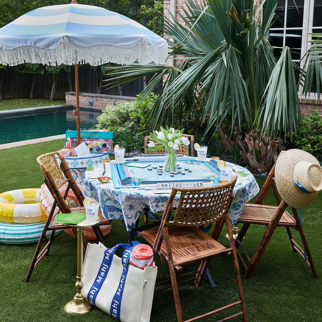 Outdoor backyard scene with a round table covered with a blue and white tablecloth, decorated with a vase of white flowers, surrounded by six wooden chairs, some with hats on them. A beach ball float is on the grass, and an umbrella provides shade. Pool in the background, trees, and plants nearby.