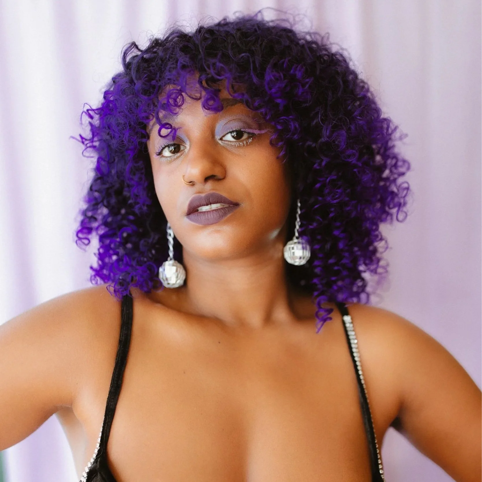 A woman with purple curly hair, wearing silver disco ball earrings and a black strap top with rhinestone details, posing against a light purple background.