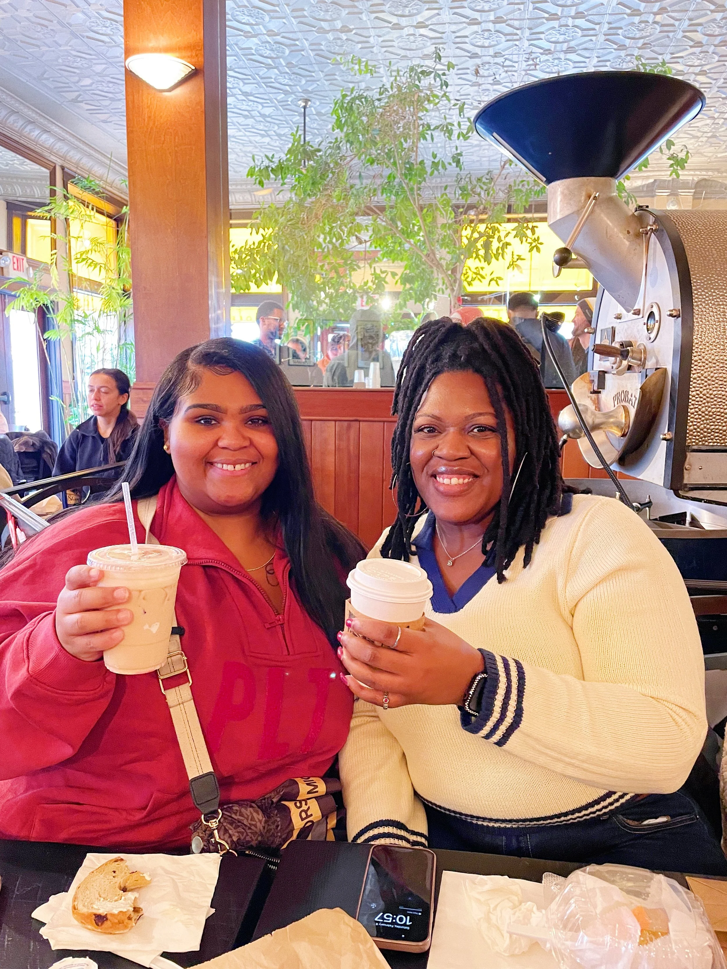 Two women smiling and holding drinks, sitting at a table in a restaurant with a coffee grinder near them.