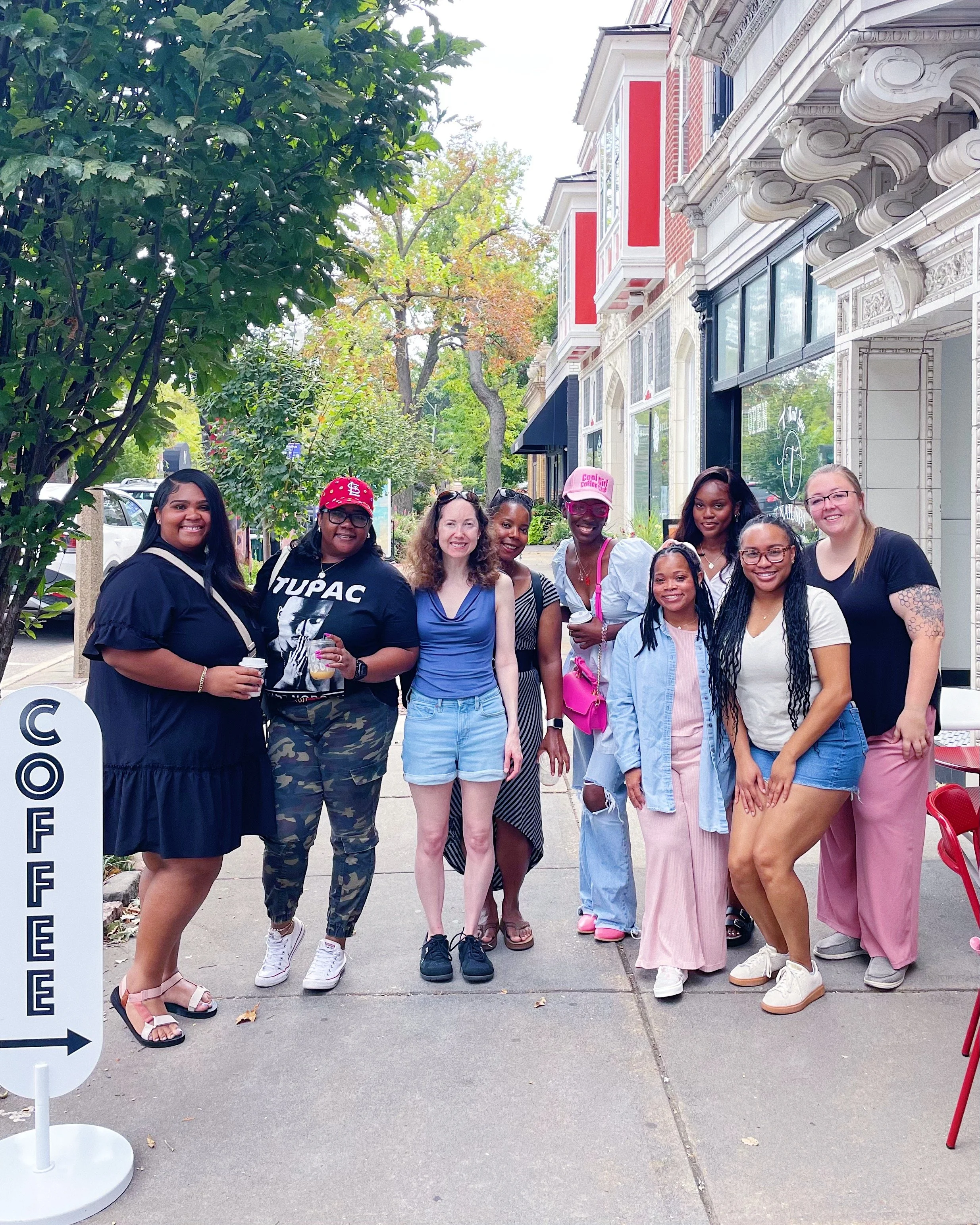 Group of women standing on a city sidewalk, smiling, with trees and storefronts in the background.