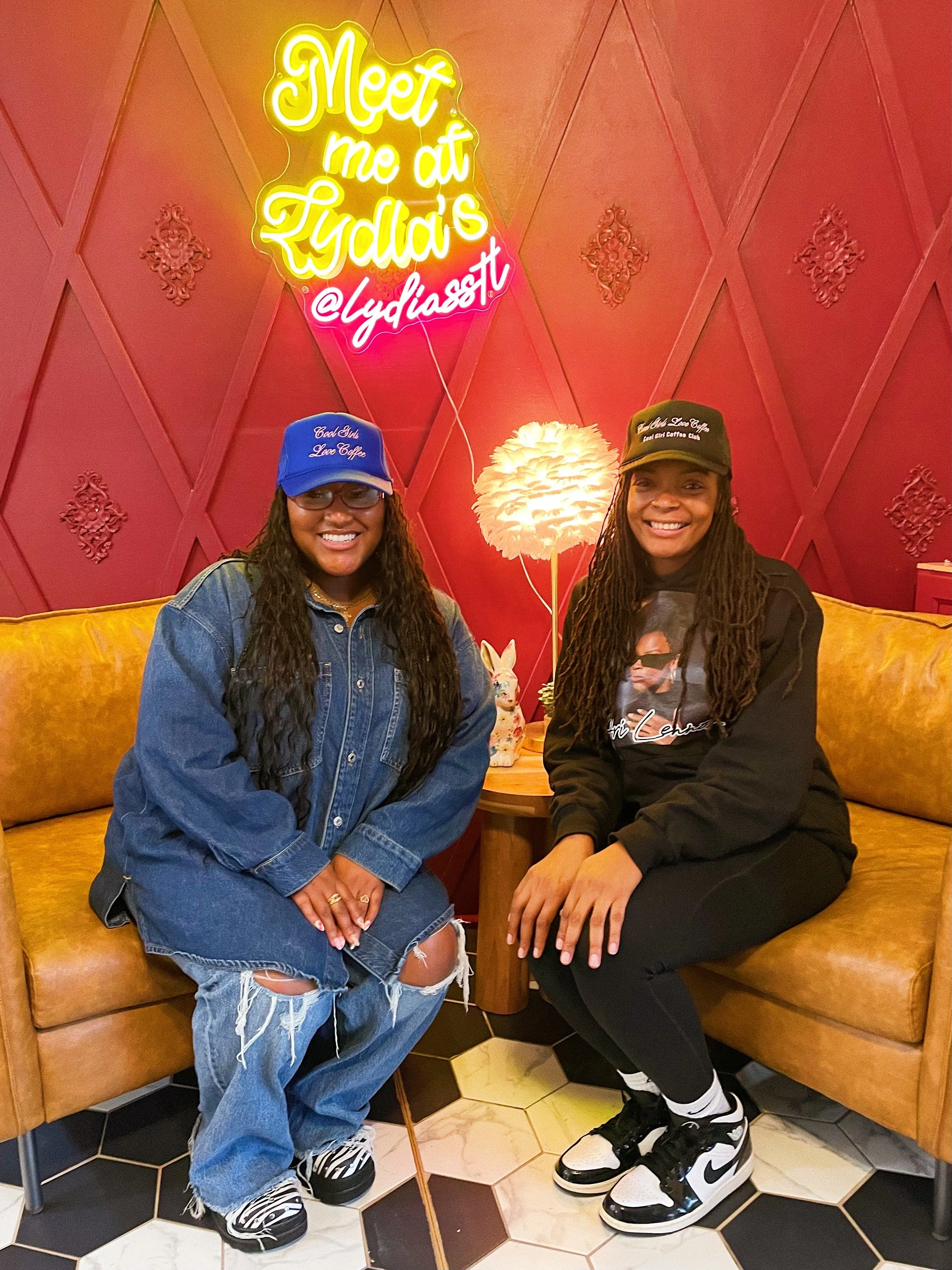 Two women sitting on a yellow couch in a cafe, smiling at the camera. The woman on the left wears a blue cap, denim jacket, ripped jeans, and sneakers. The woman on the right wears a black hoodie, black leggings, and Nike sneakers. Behind them is a r