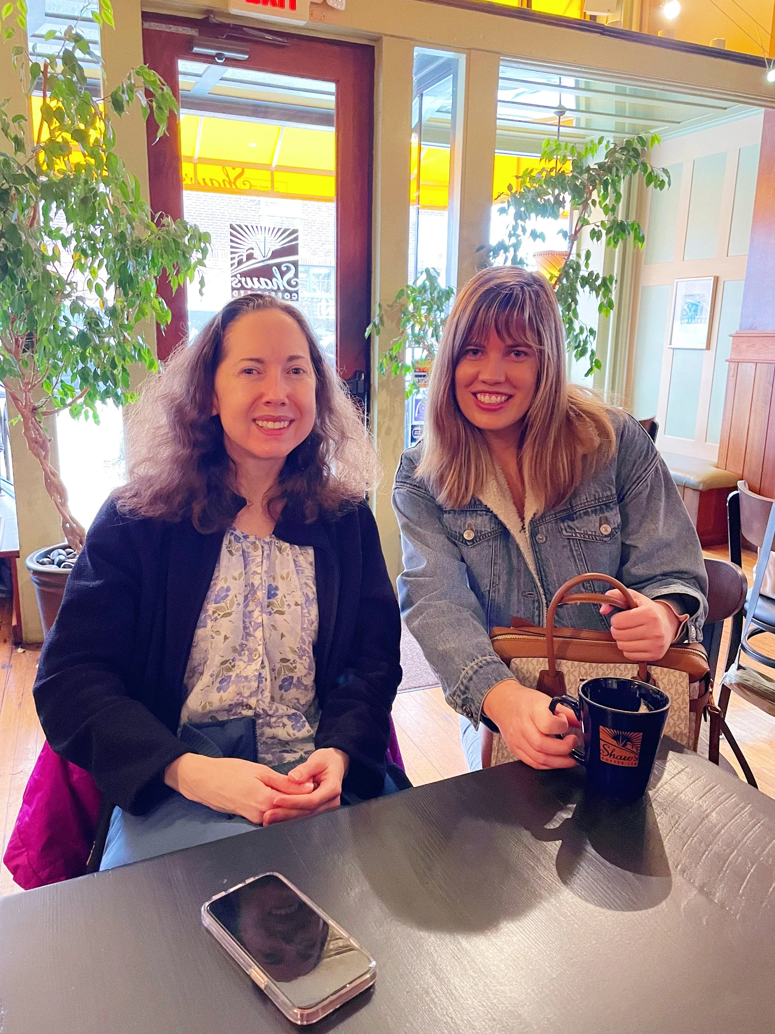 Two women sitting at a table inside a coffee shop, smiling at the camera. One has curly hair and is wearing a black jacket, the other has straight hair and is wearing a denim jacket. There are potted plants behind them and a window with a yellow awni