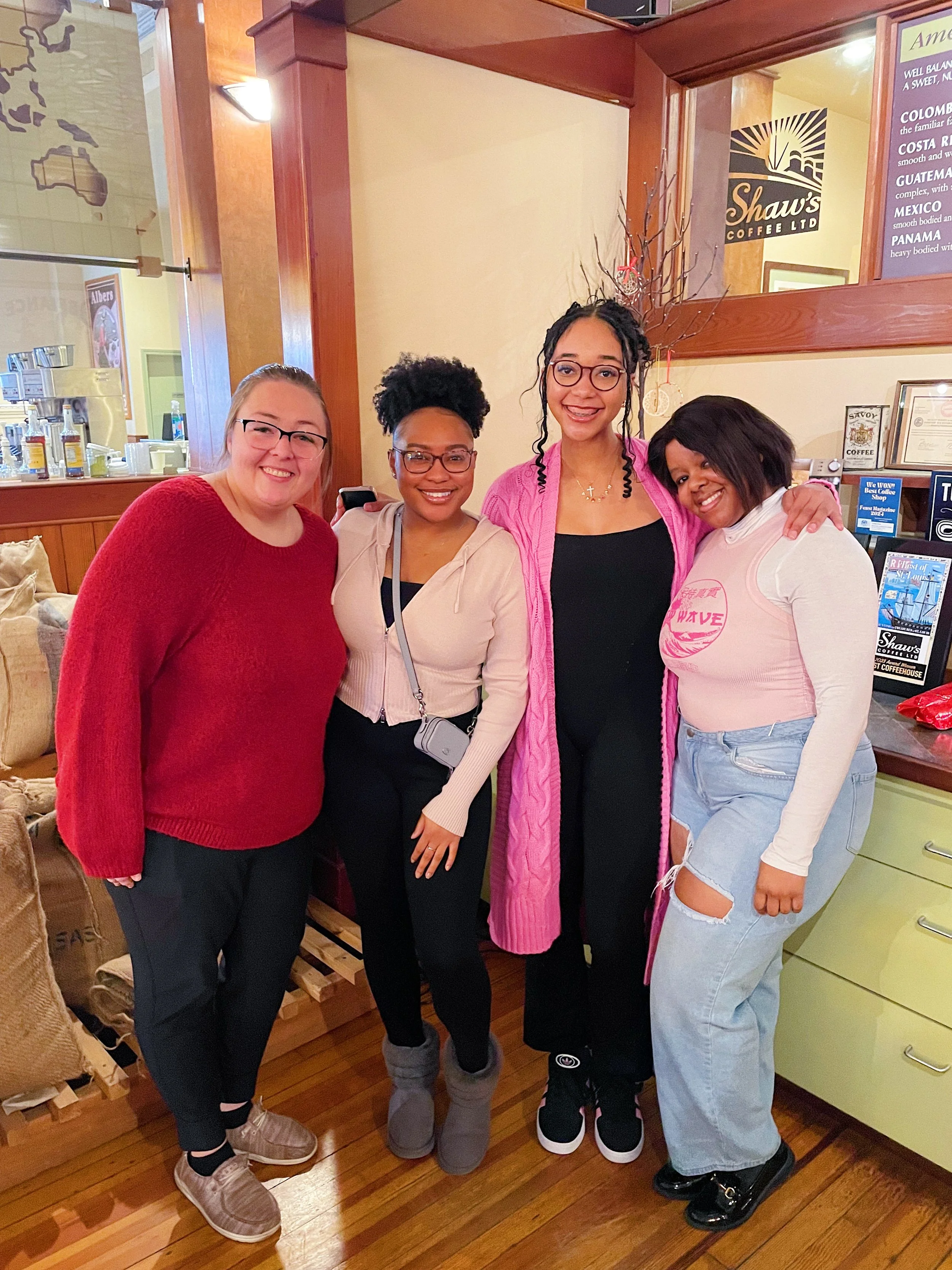 Four women smiling and standing close together inside a cozy cafe, with a wooden counter, coffee machines, and food items in the background. They are dressed casually and appear happy.