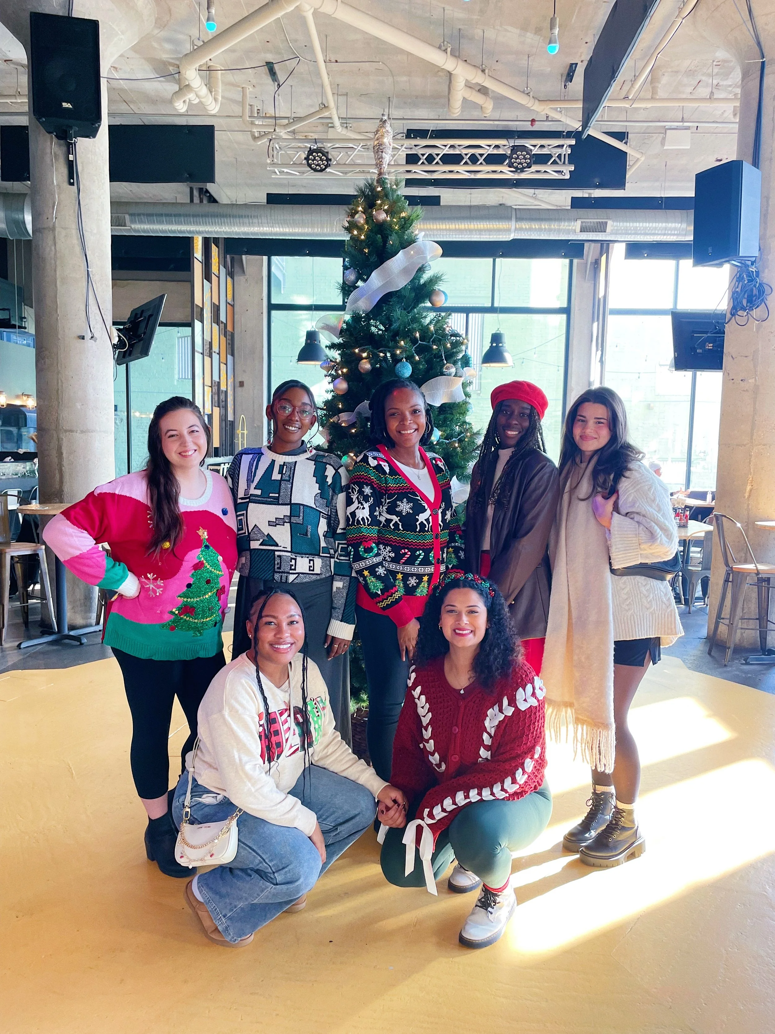 A group of seven diverse women in festive holiday sweaters and accessories, standing and kneeling in front of a Christmas tree with ornaments and ribbon, inside a modern, industrial-style space with large windows.
