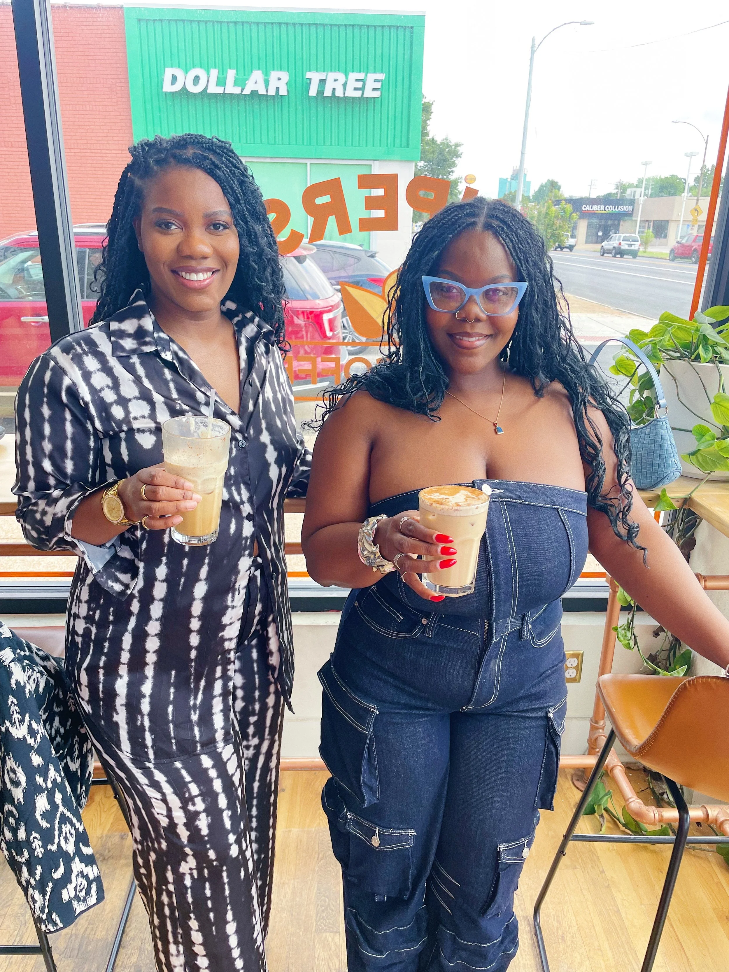 Two women are standing inside a cafe, holding drinks. One woman is wearing a black and white patterned jumpsuit and glasses. The other woman is wearing a strapless denim jumpsuit and blue glasses. There is a large window behind them with the words 'D