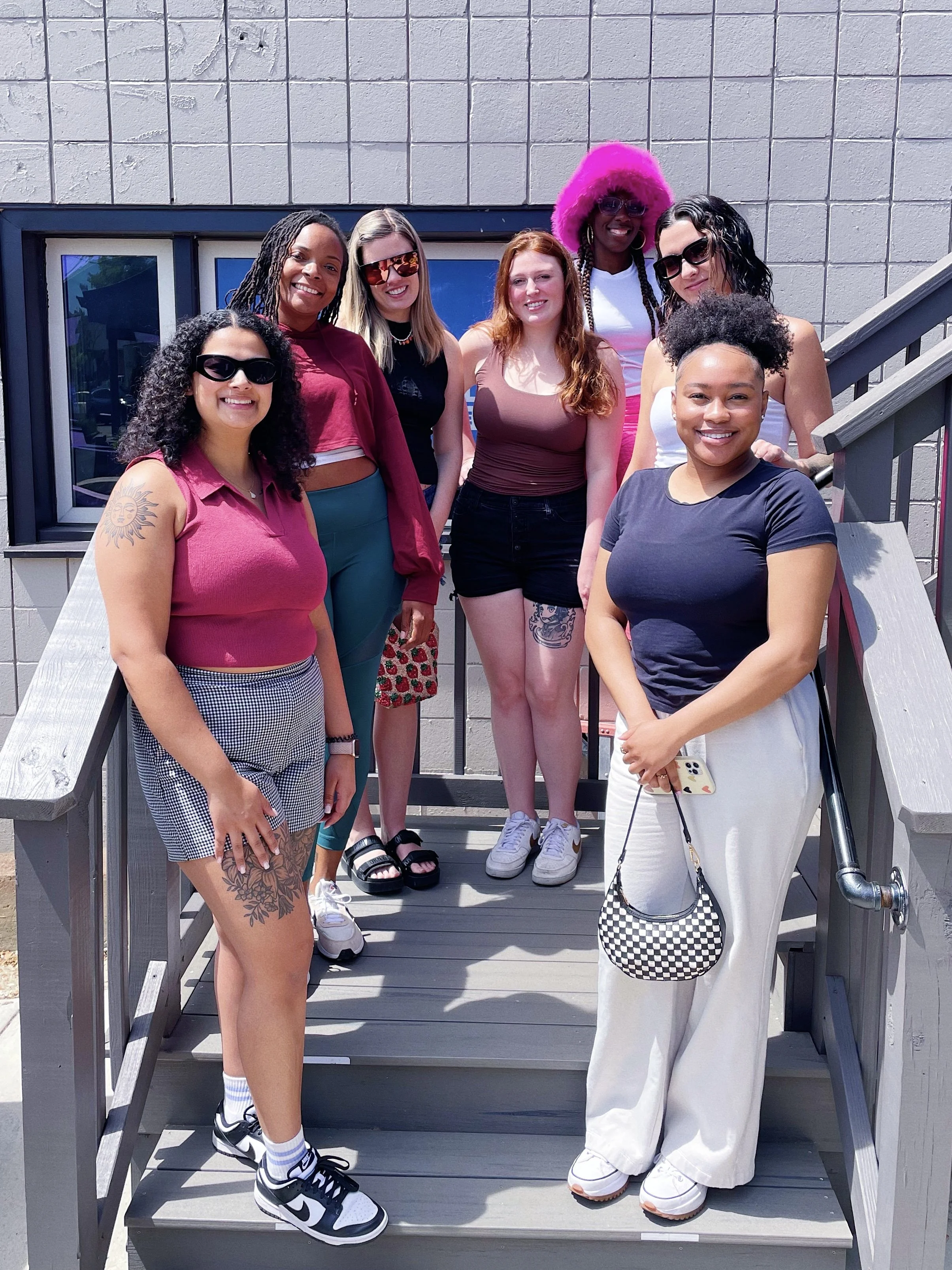 Group of nine women standing on a wooden staircase outside a building, smiling for a photo. They are dressed in casual summer clothing and sunglasses, with diverse hairstyles and skin tones.
