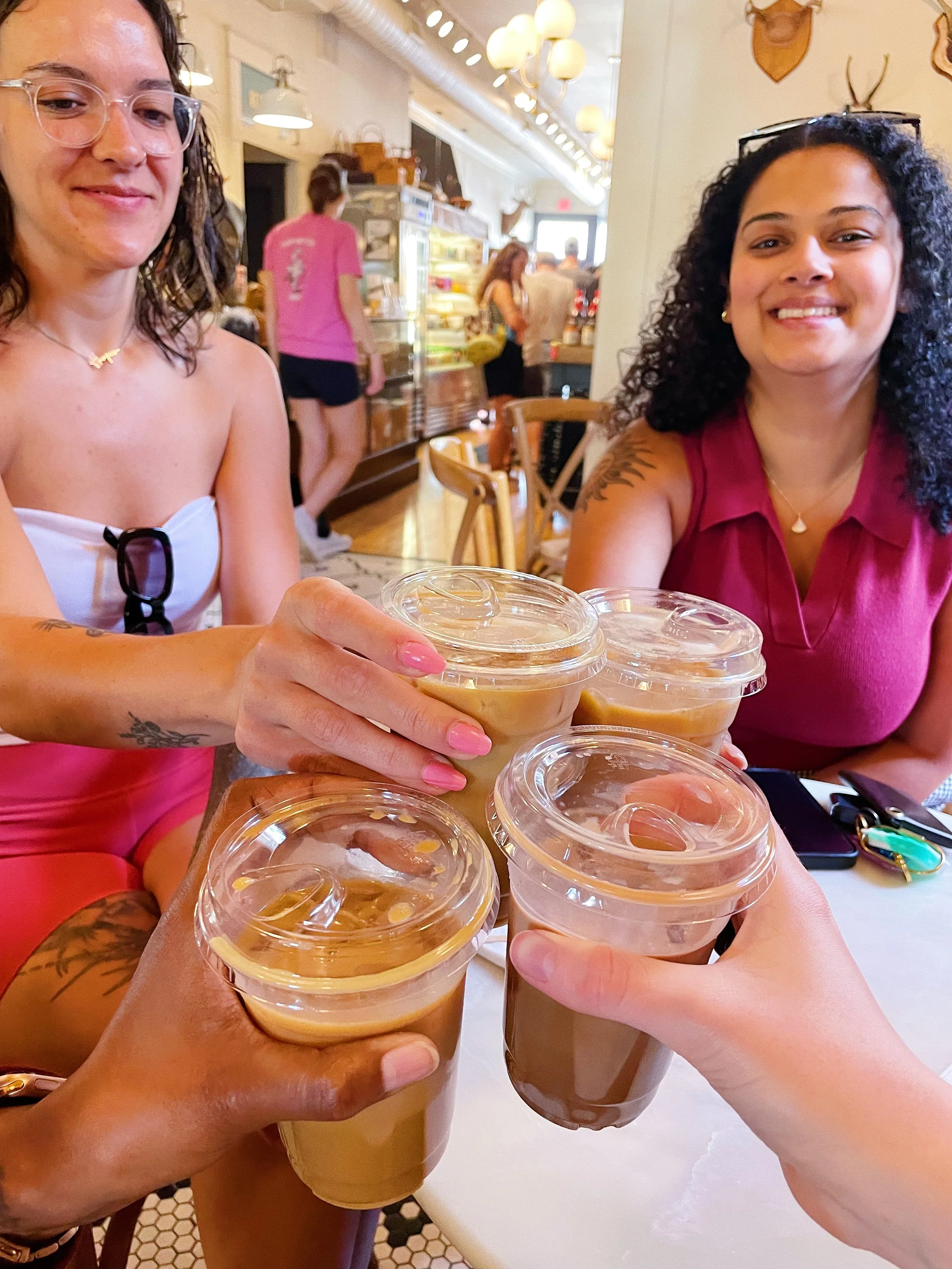 Four people toasting with plastic cups of iced coffee or iced chocolate at a restaurant or cafe, with two women sitting at a table and clinking their cups together.