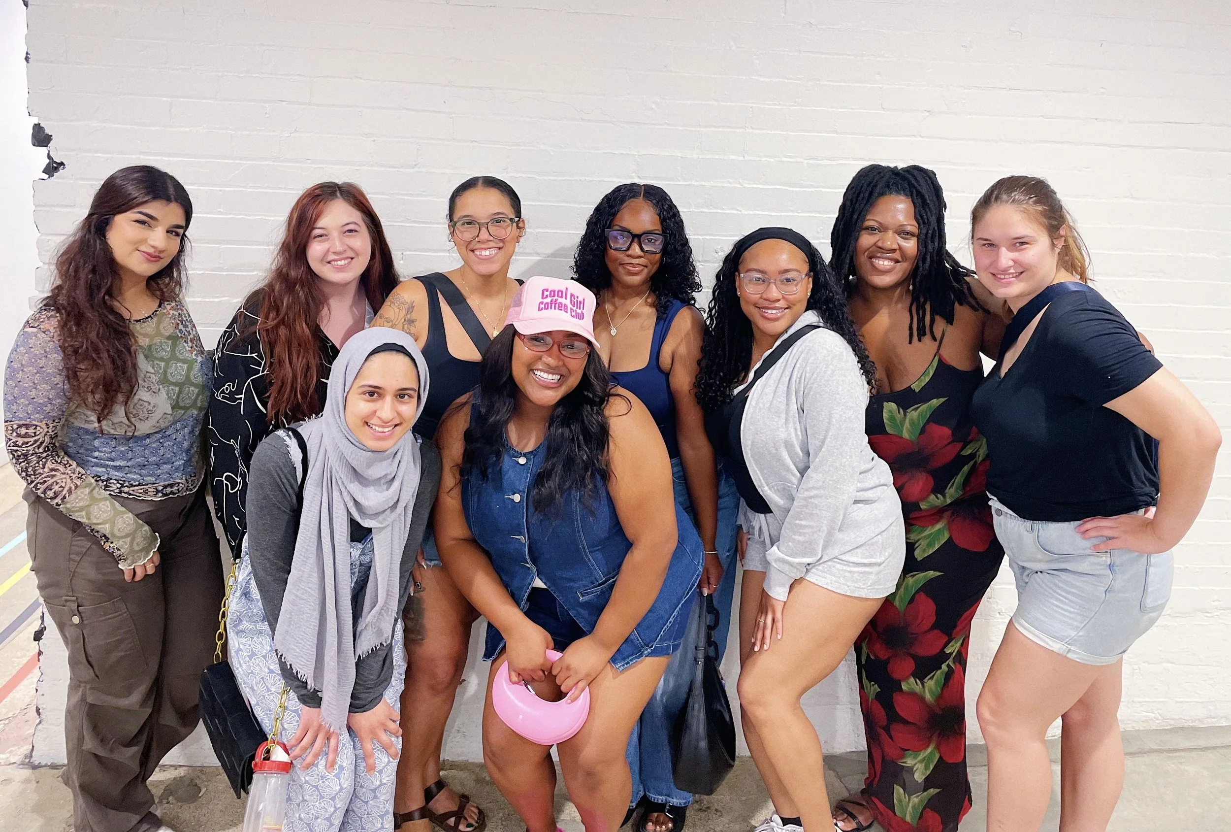 Group of ten diverse women standing together against a white brick wall, smiling at the camera, dressed casually.